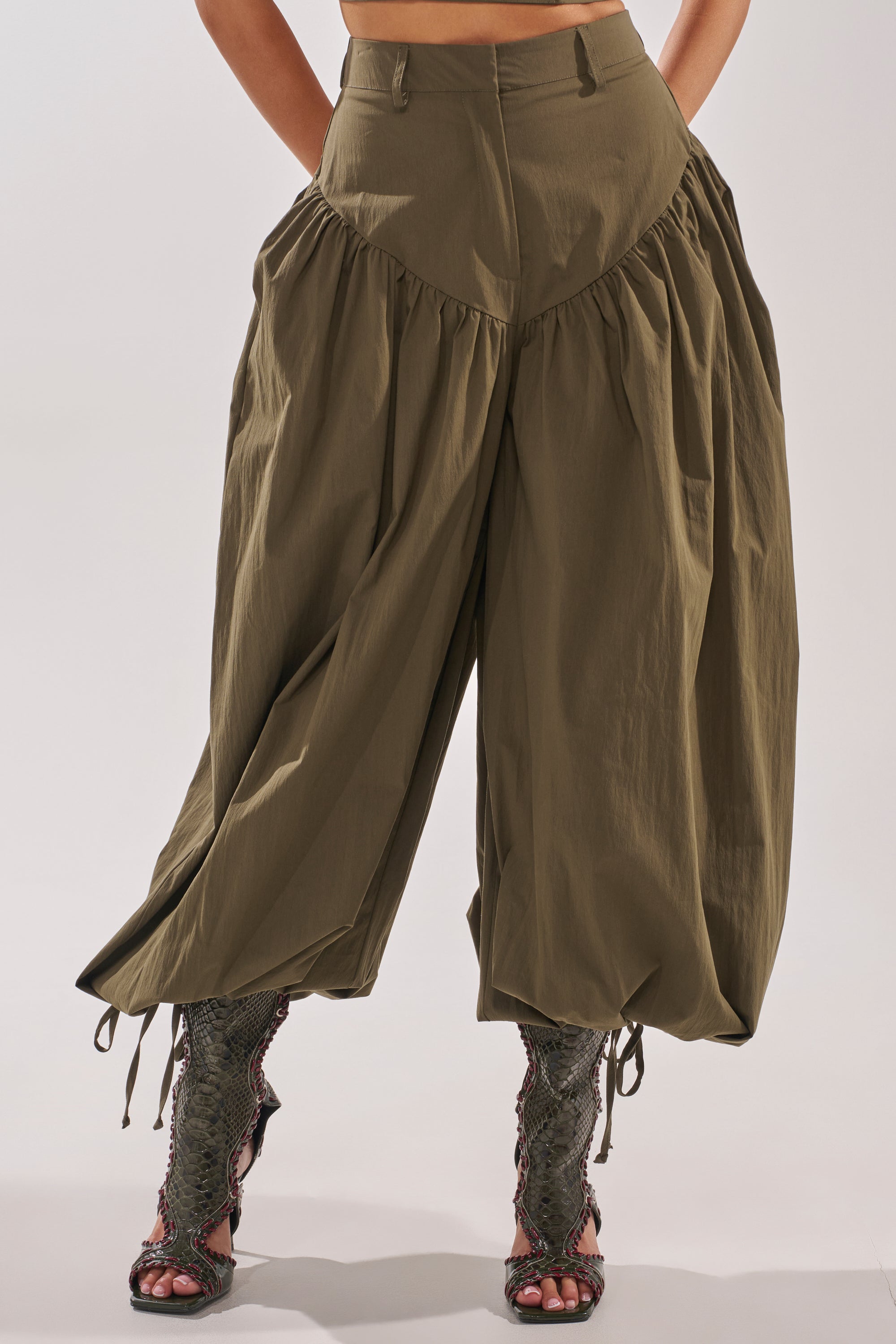 A person models the LUCKY FOR YOU BALLOON PANT—high-waisted, wide-leg olive green pants with dramatic ruffles and gathered details—styled with knee-high, textured open-toe boots. The upper body is not visible. 2