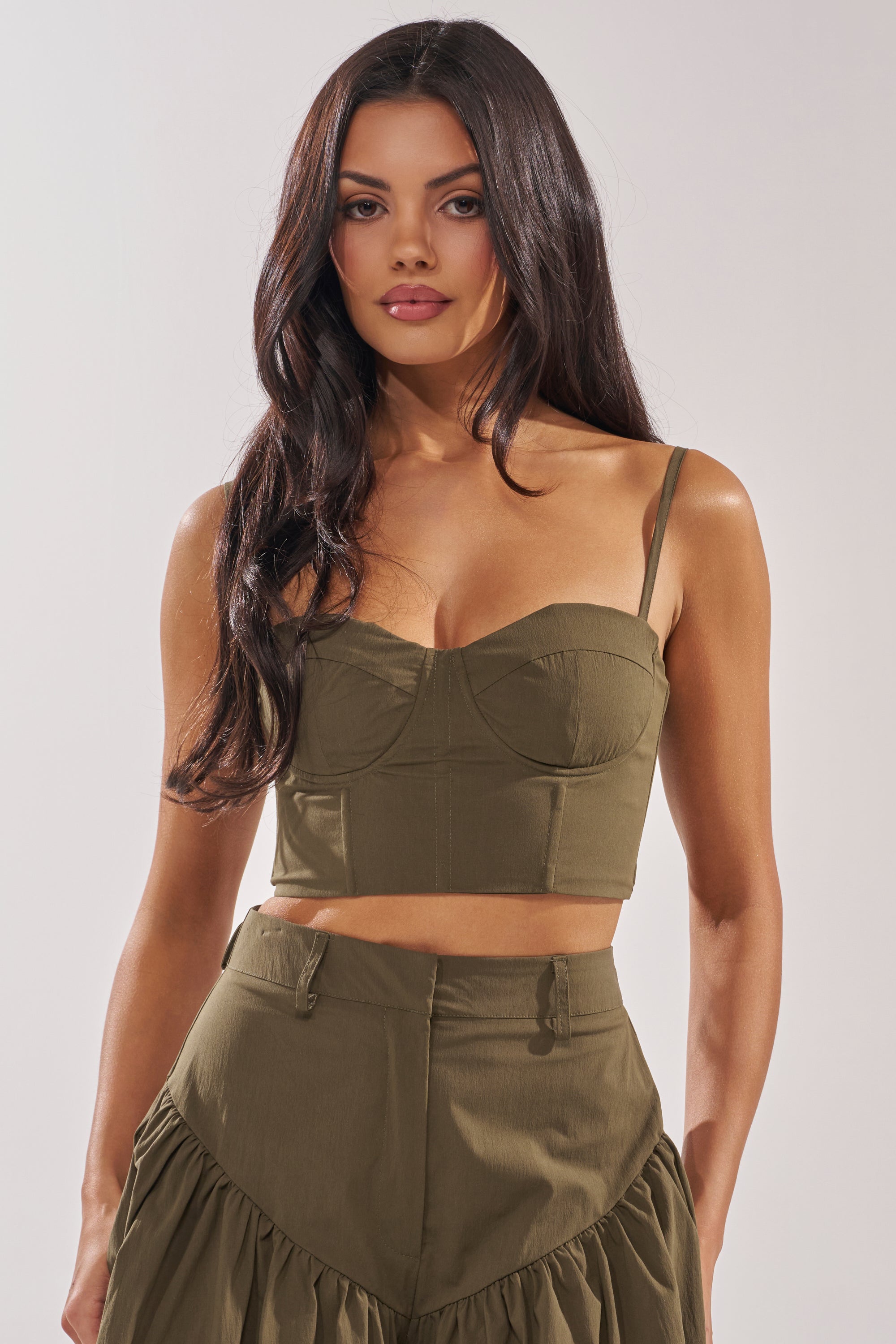 A woman with long dark hair wears the LUCKY FOR YOU CORSET, an olive green crop top, paired with a matching high-waisted skirt featuring ruffled details. She stands confidently against a plain light background. 0