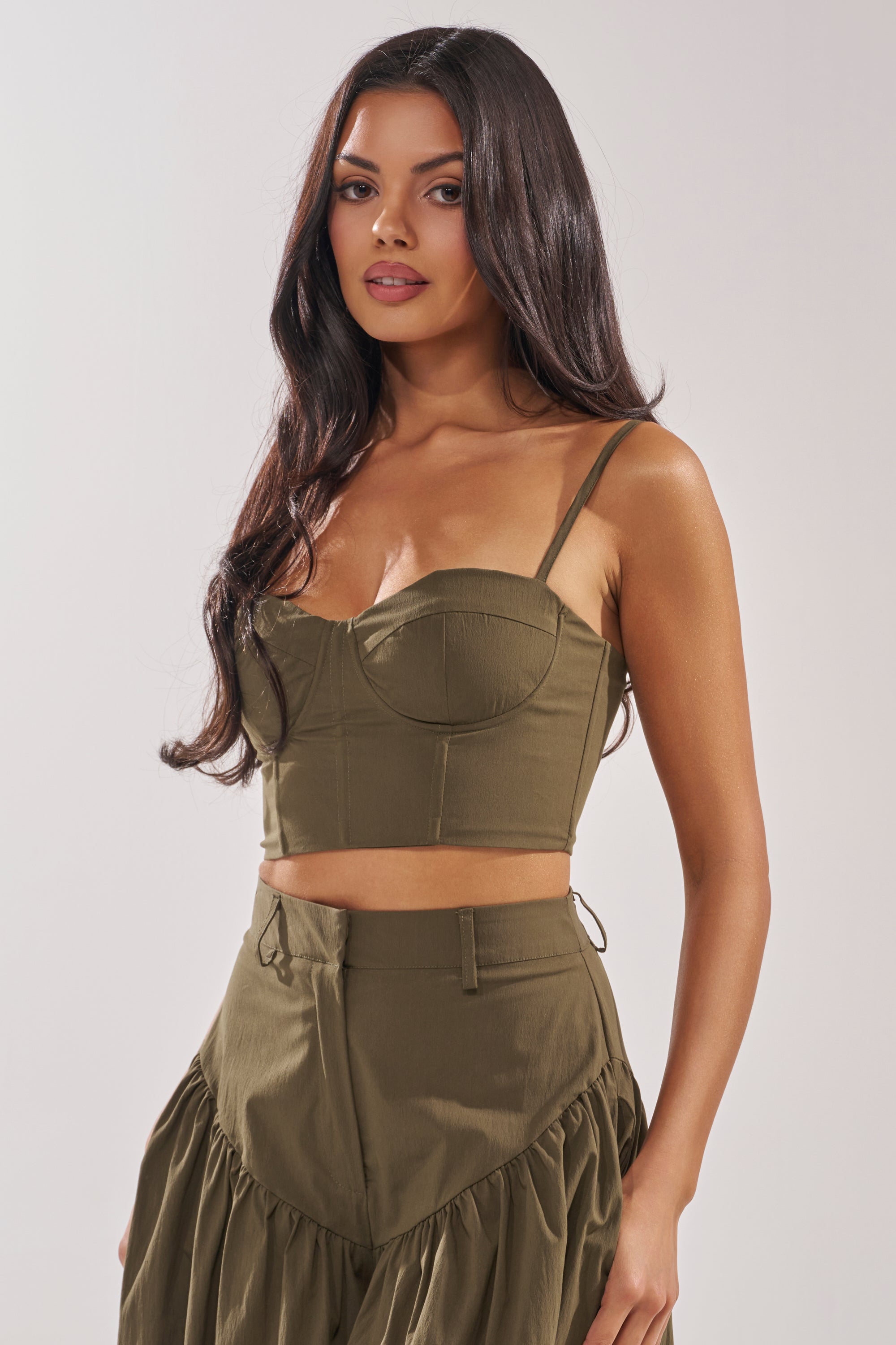 A woman with long dark hair wears the LUCKY FOR YOU CORSET in olive green with thin straps, paired with a matching high-waisted skirt with ruffle detail, standing against a plain light background. 2
