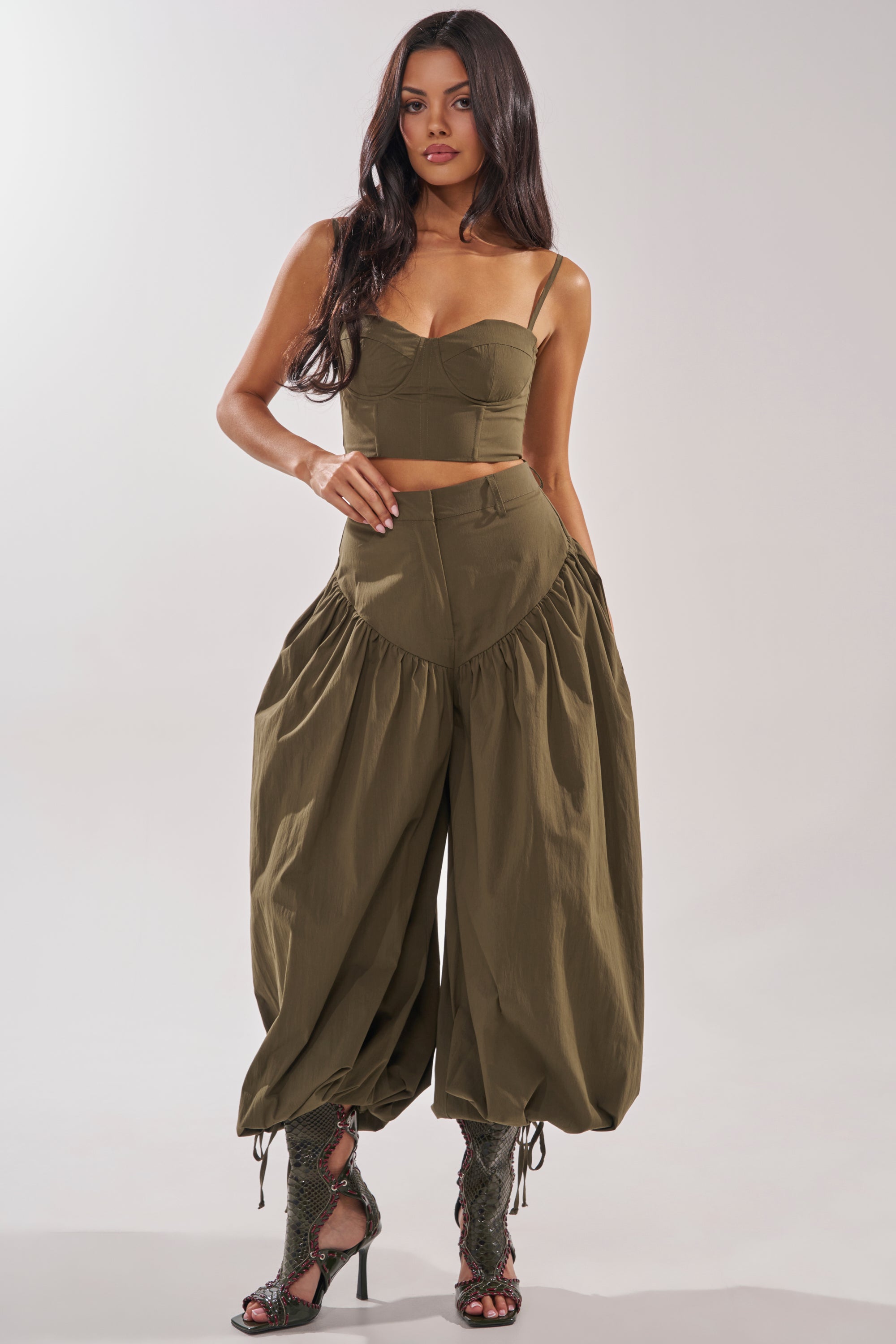 A woman poses confidently against a light background in the LUCKY FOR YOU CORSET, an olive green crop top with a sweetheart neckline, paired with matching wide-leg balloon pants and lace-up high heel sandals. Her long dark hair is loose. 6