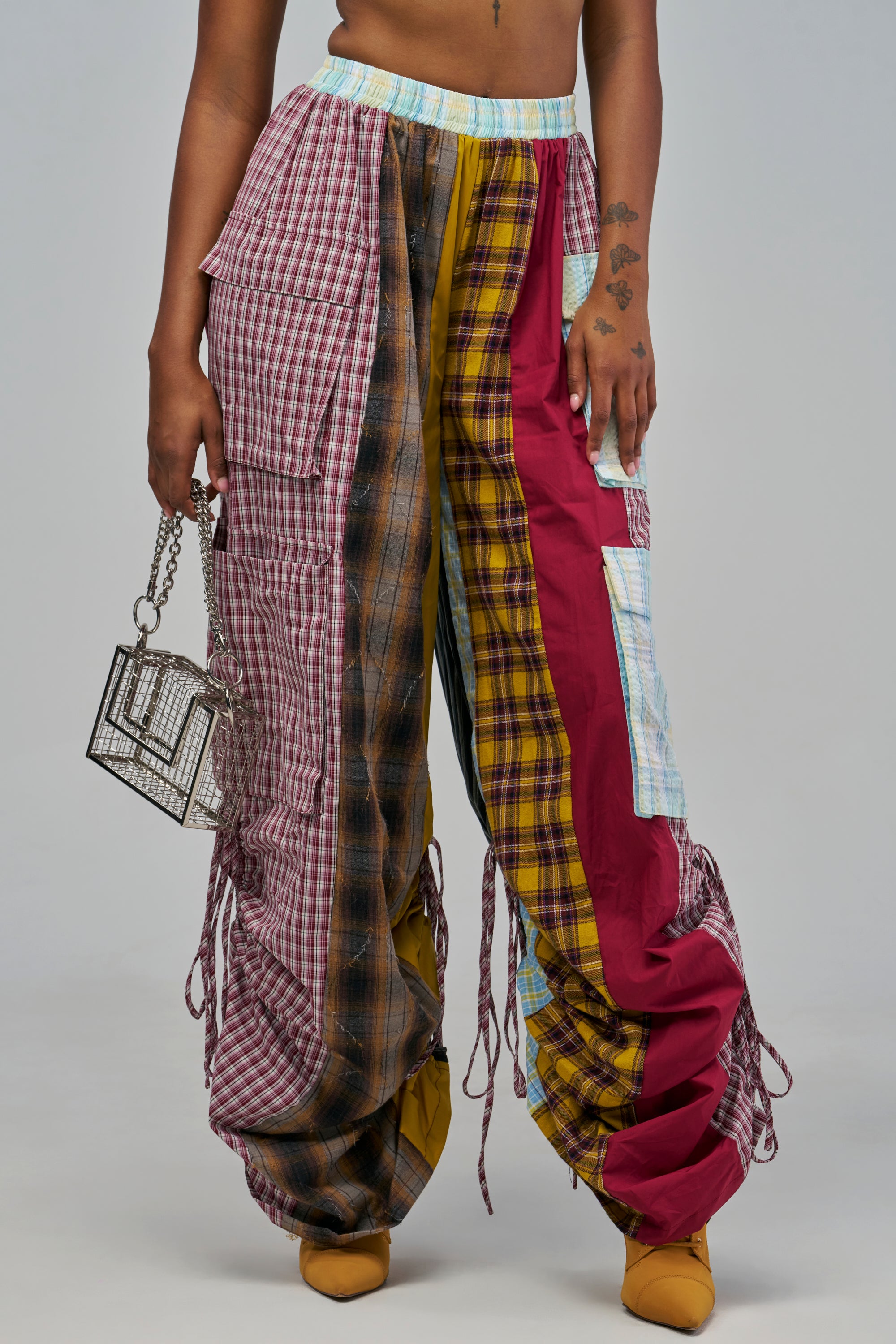 MAIN MOMENT PLAID PANT: high-waisted, wide-leg cargo pants featuring a patchwork of red, yellow, and brown plaid patterns, solid red panels, and drawstrings at the hem, worn with a small metallic cage handbag.