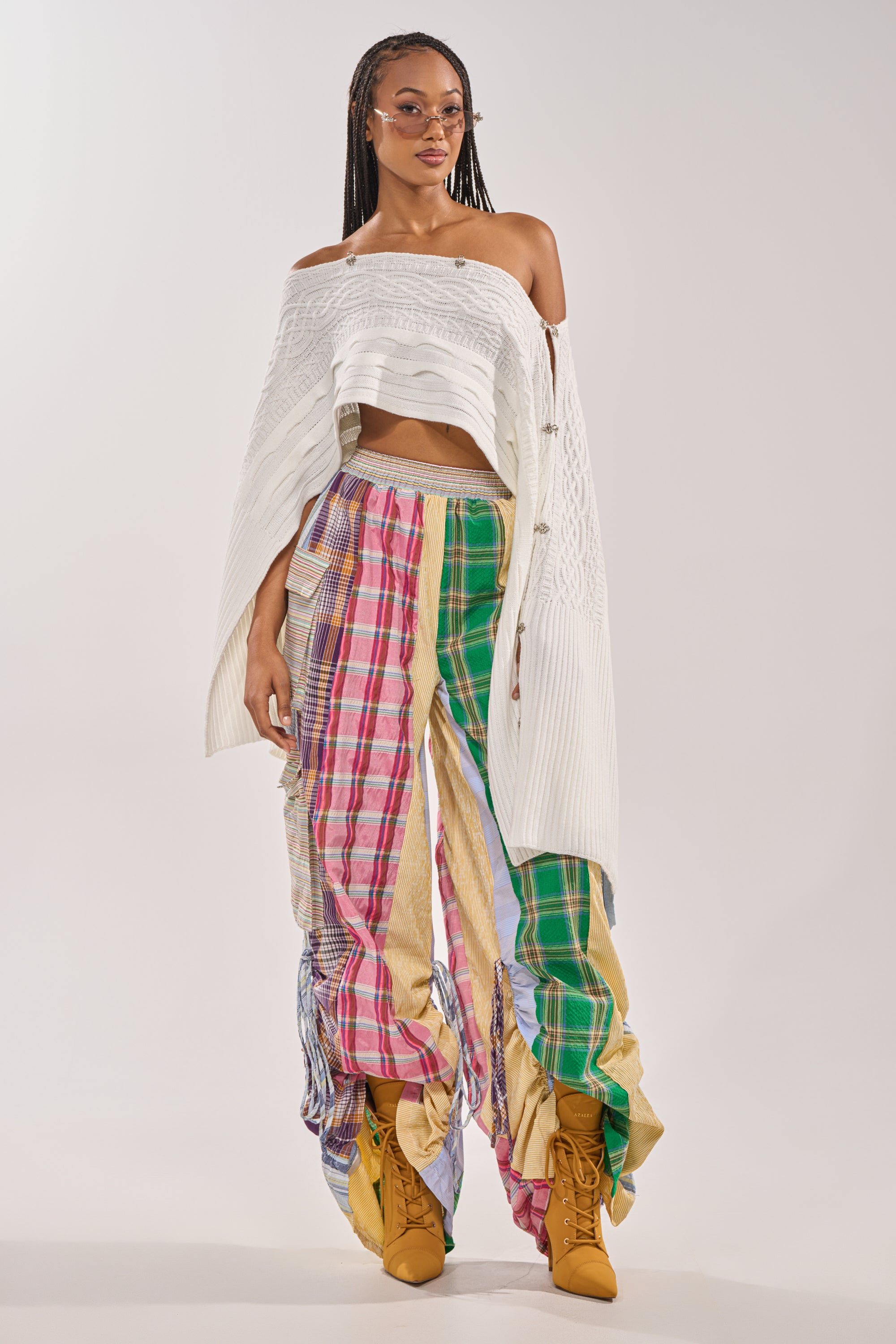A woman stands confidently in MIXED PLAIDS JOGGERs, a white off-the-shoulder top, mustard yellow lace-up boots, and pink-tinted sunglasses, embodying bold streetwear style against a plain background. 3