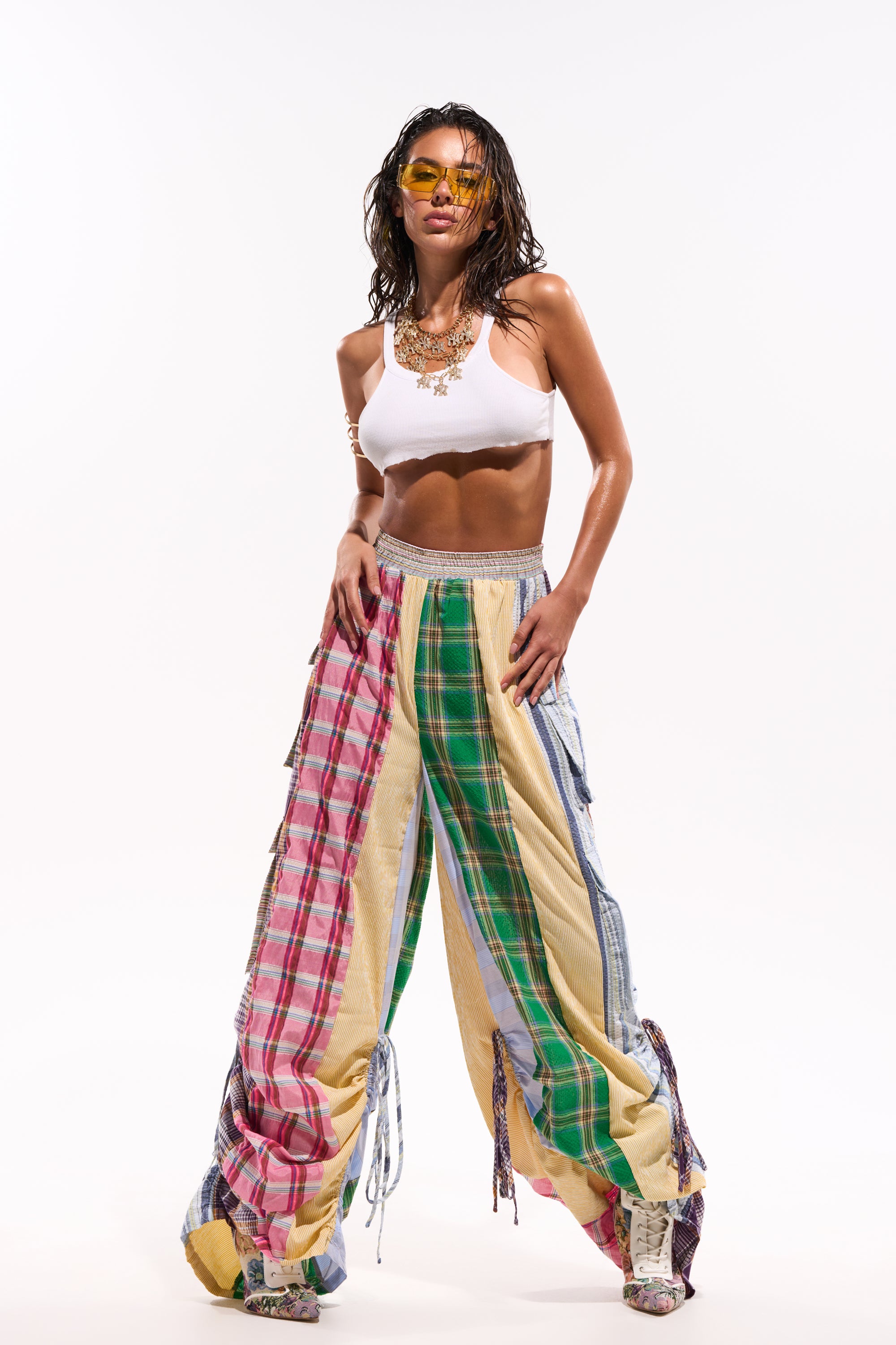 A woman stands confidently against a white background wearing MIXED PLAIDS JOGGER pants, a white crop top, chunky shoes, layered necklaces, and yellow sunglasses. 2