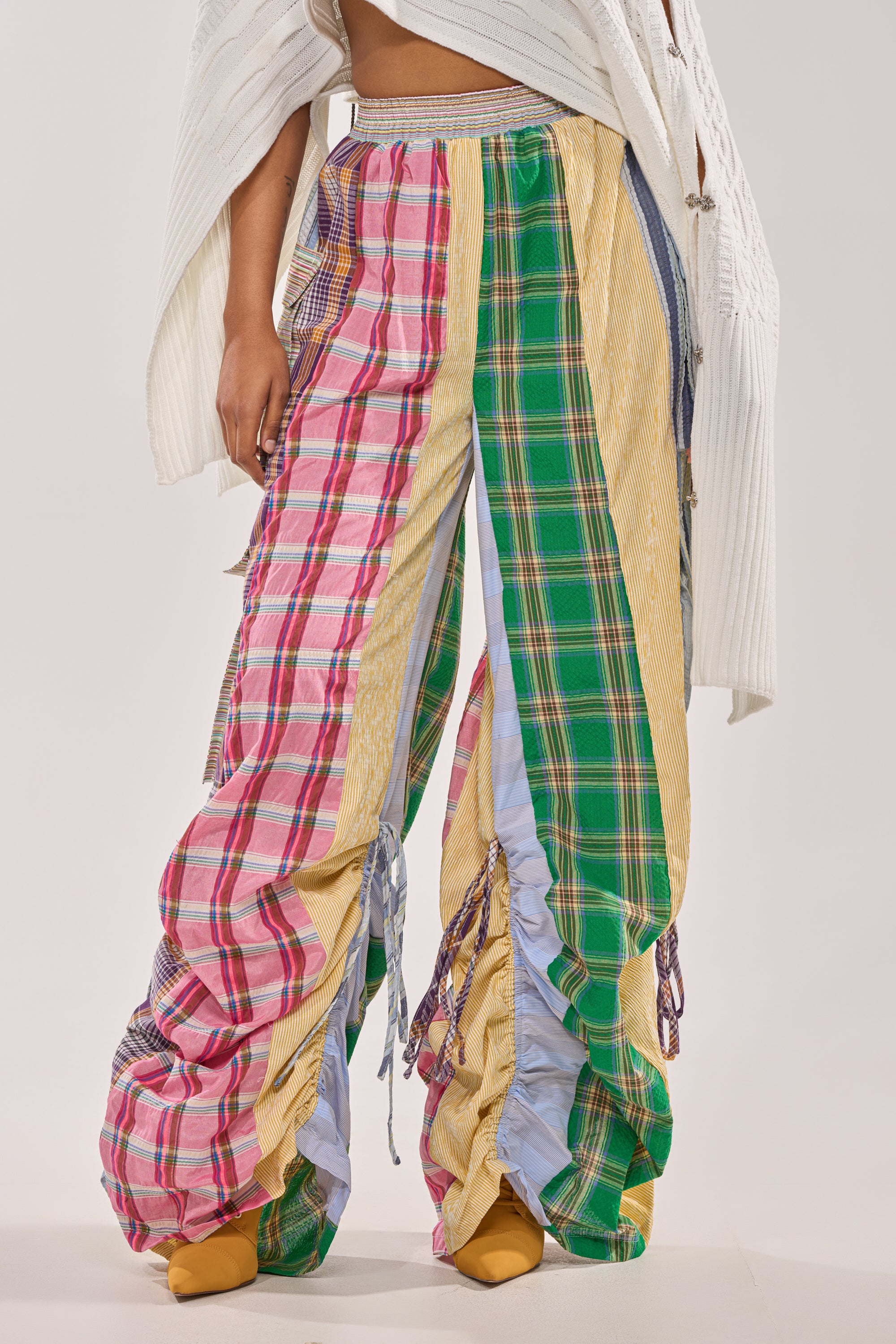 A person wears the MIXED PLAIDS JOGGER—loose cargo joggers in pink, green, yellow, and blue plaid—with a white textured sweater and mustard-yellow shoes, standing against a plain background. 1