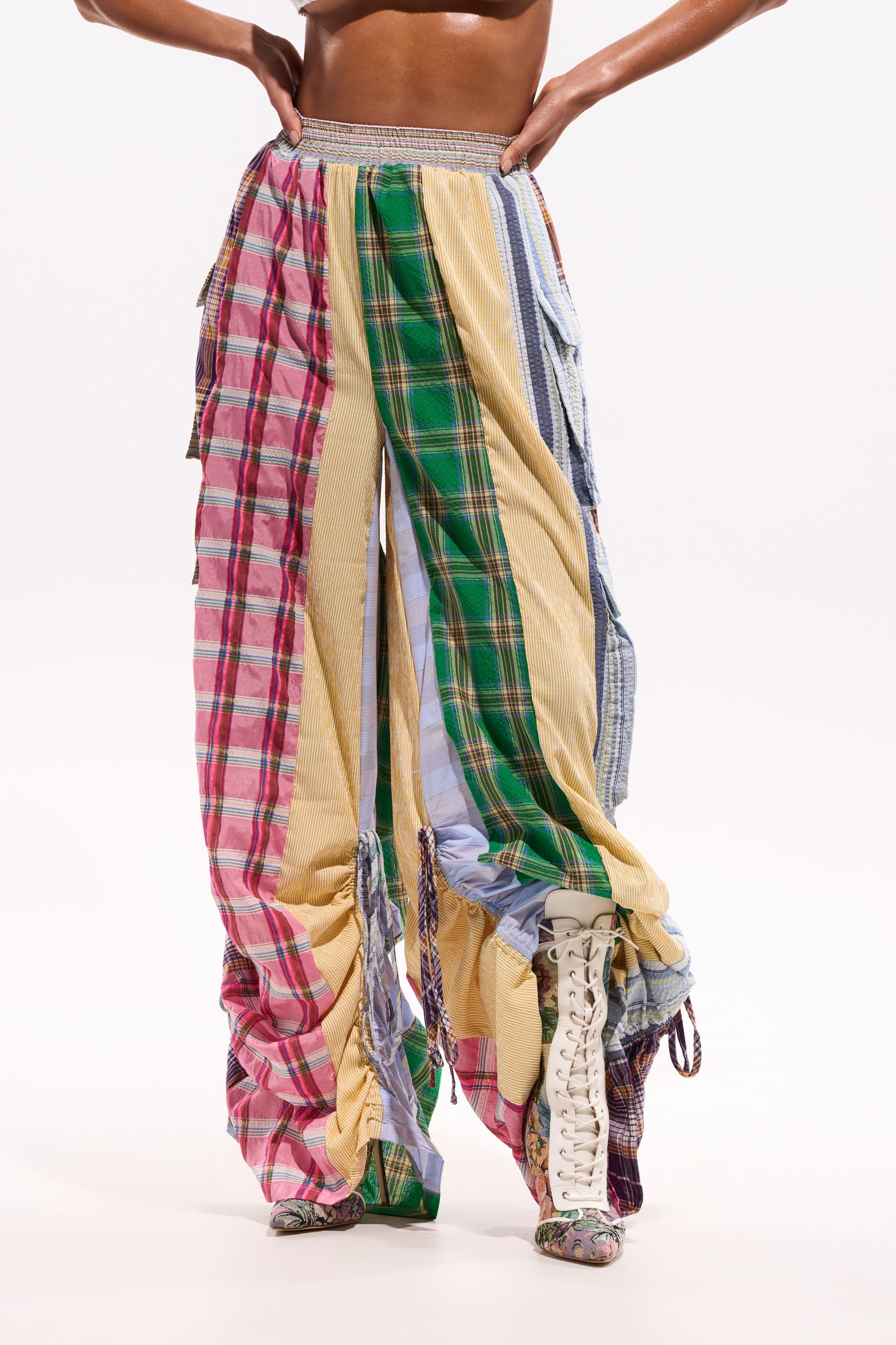 Someone wearing the MIXED PLAIDS JOGGER, featuring wide-leg patchwork cargo style with colorful plaid and striped patterns, gathered ruching, and paired with white lace-up boots; their hands rest on their hips. 0