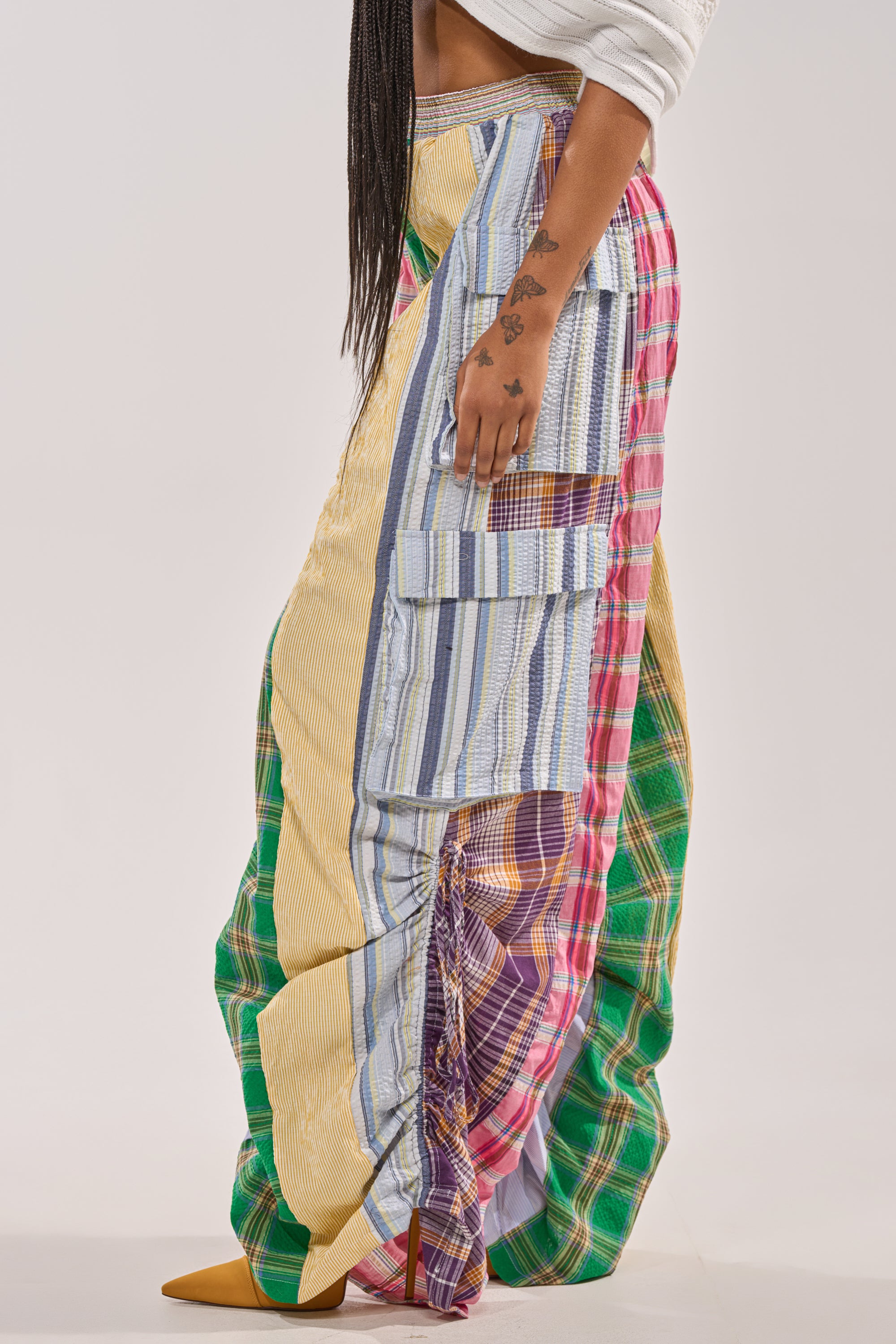 Wearing the MIXED PLAIDS JOGGER with colorful plaid and striped patches, a large side pocket, and yellow pointed shoes, this person’s streetwear look pops against a plain background. Small tattoos decorate their hand for extra edge. 5