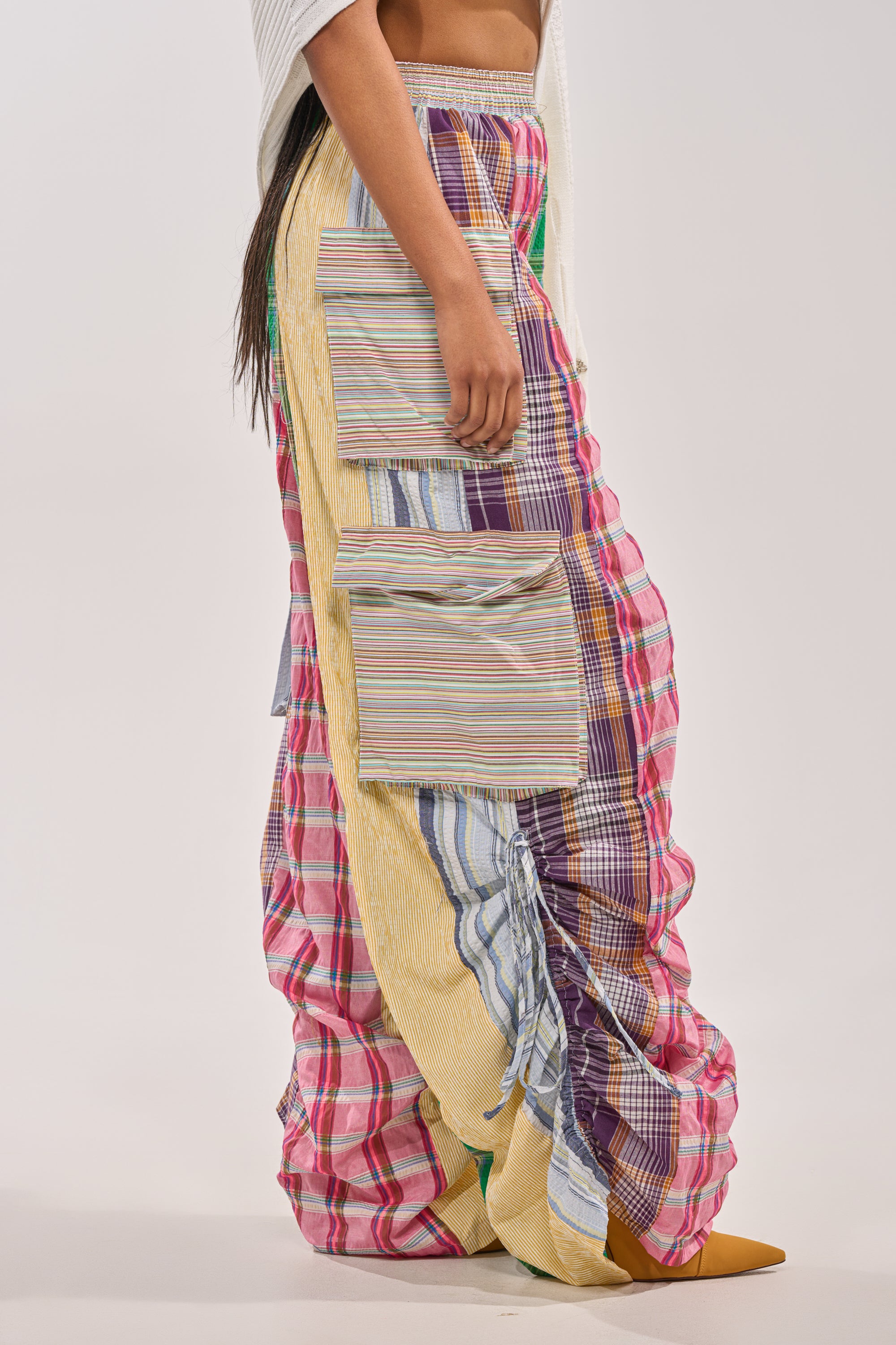 A person models the MIXED PLAIDS JOGGER, featuring bold patchwork cargo joggers with plaid and striped patterns and large striped side pockets. The image is cropped to the lower body against a neutral background, showcasing standout streetwear style. 6