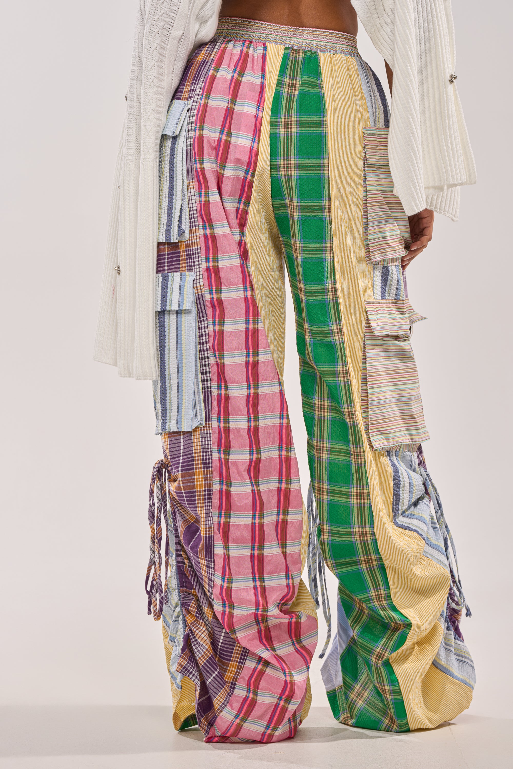 A person wears the MIXED PLAIDS JOGGER, featuring vibrant plaid and striped patchwork, paired with a textured white top. The streetwear-inspired outfit highlights bold patterns and mixed textures. 7
