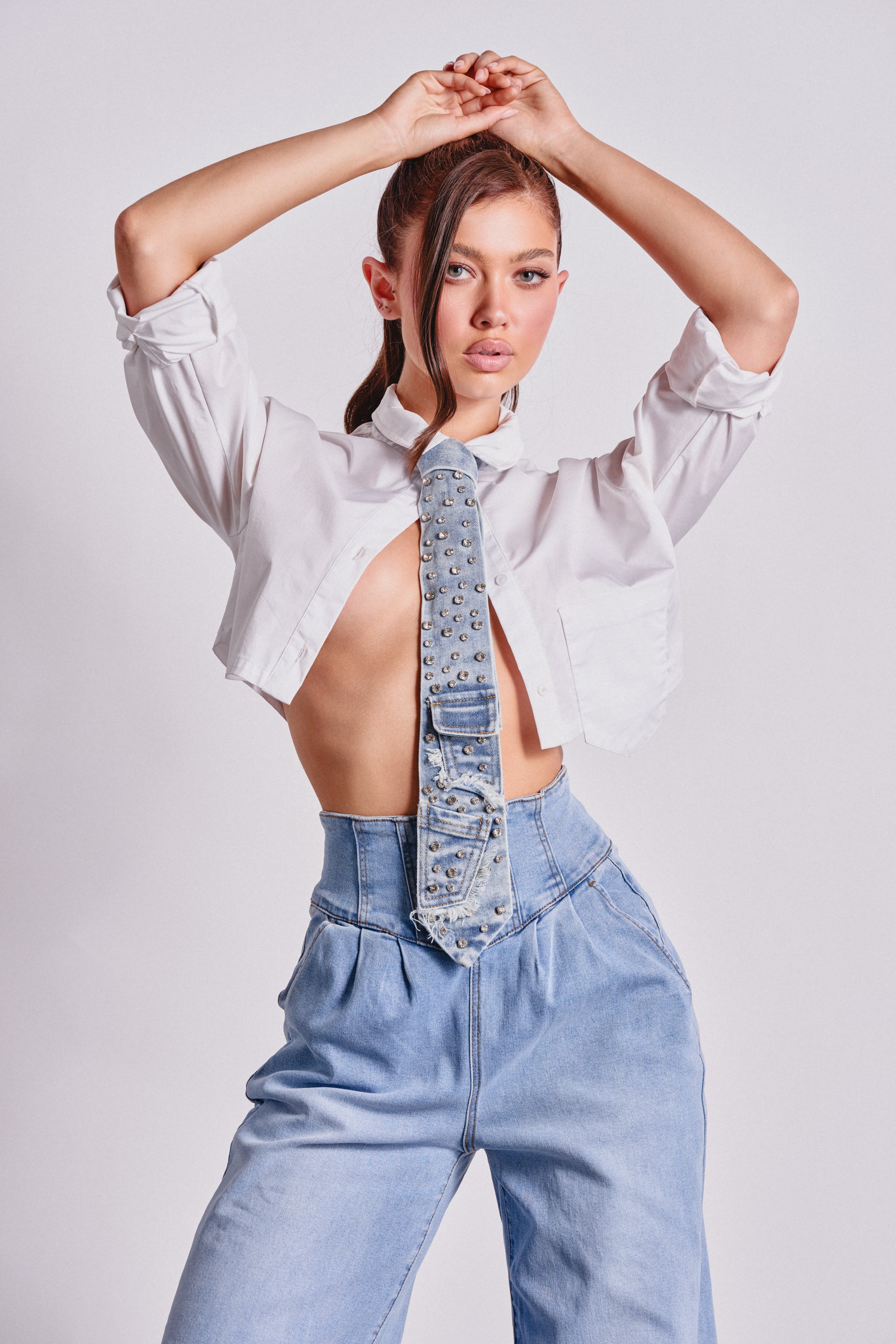 A woman poses with her arms raised, wearing a cropped white shirt, the DENIM AND DIAMONDS TIE, and high-waisted light blue cargo jeans against a plain light background. 1