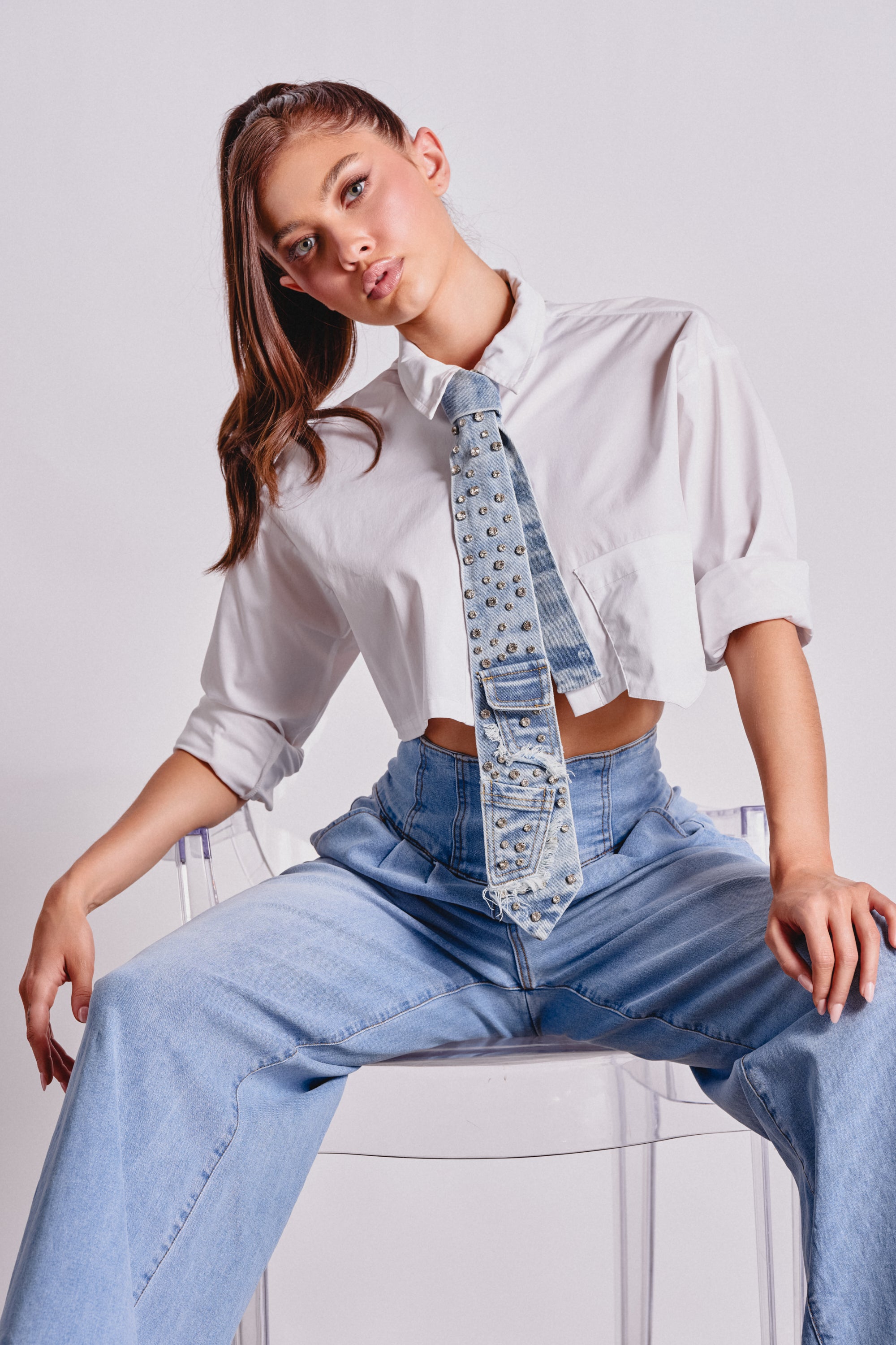 A young woman in a white cropped shirt and light blue wide-leg jeans sits on a clear chair, her hair in a high ponytail. She wears the DENIM AND DIAMONDS TIE, making it the standout statement accessory of her look. 2
