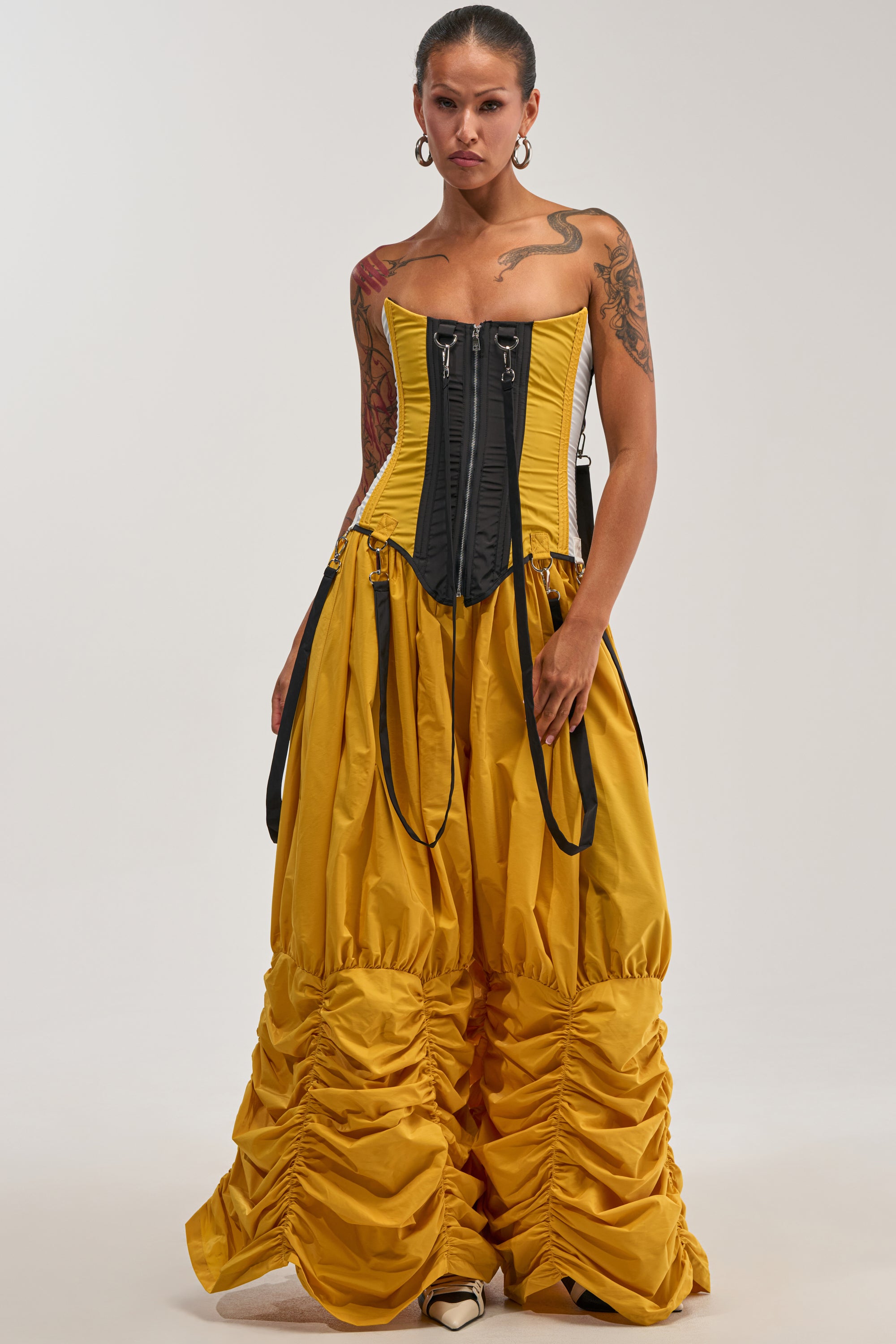 A person stands wearing the DON'T MIND ME RUCHED JOGGER IN YELLOW with a black corset top and tattooed arms, posing against a plain background, effortlessly channeling the vibrant energy of festival style.