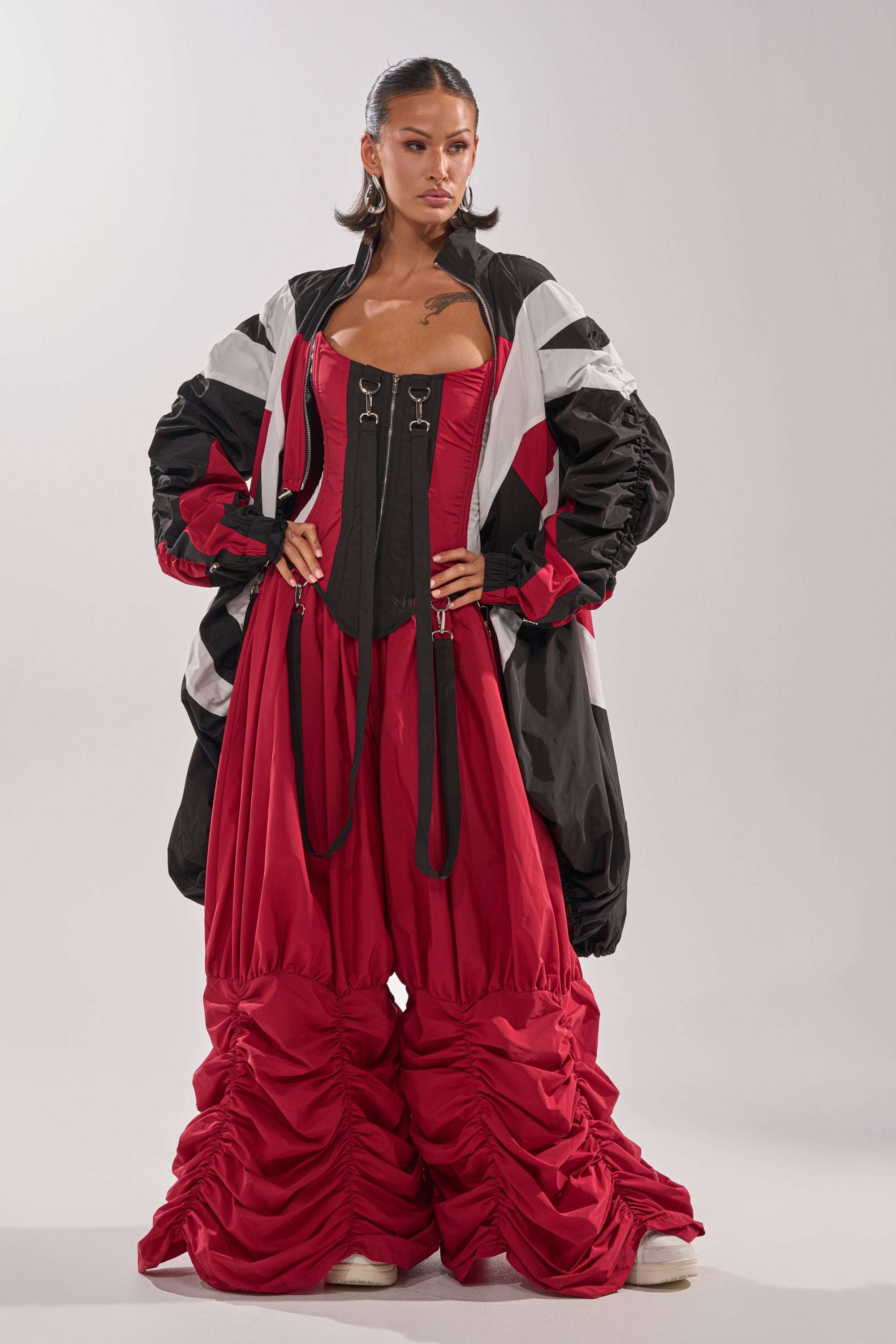A woman stands confidently in a dramatic look featuring the DON'T MIND ME STACKED JOGGER, paired with a corset top, red windbreaker, and oversized puffy jacket. She sports slicked-back hair, bold earrings, and visible tattoos. 2