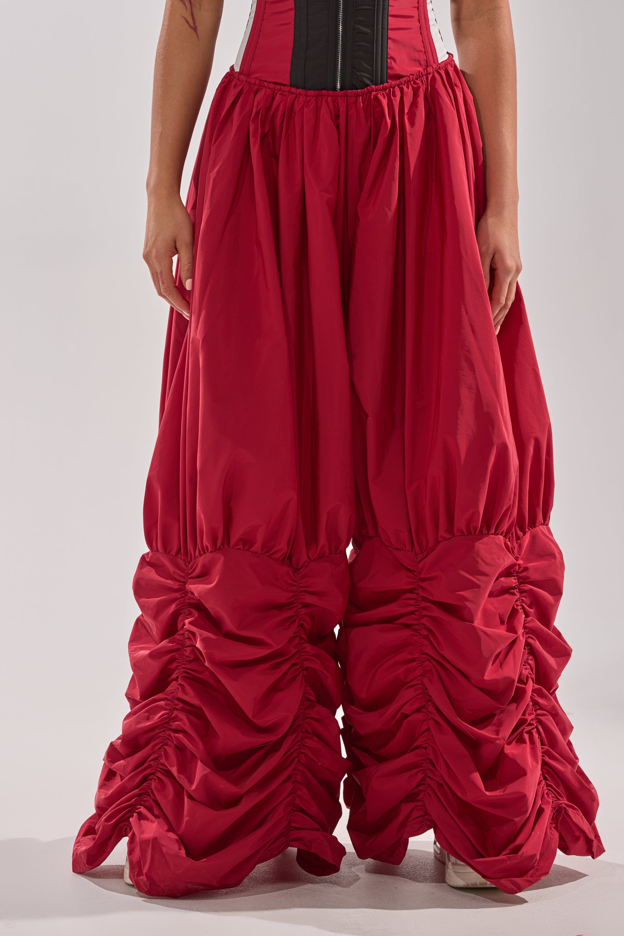 A person wears DON'T MIND ME STACKED JOGGER pants in red with ruched, voluminous style and a fitted black and white corset top, standing against a plain light background. Only the lower torso and legs are visible. 3