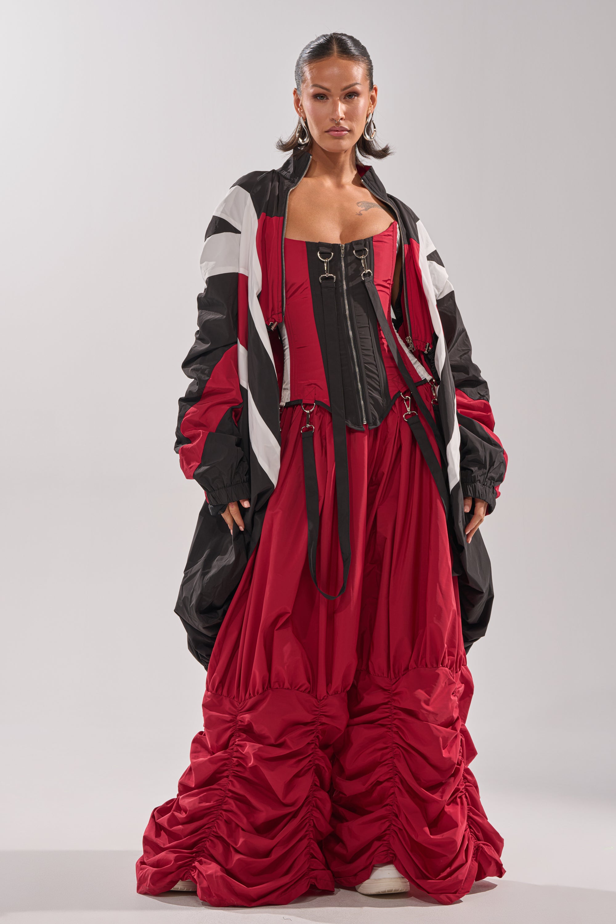 A woman stands confidently in the DON'T MIND ME STACKED JOGGER, paired with a black corset-style top and an oversized red windbreaker, accessorized with hoop earrings and slicked-back hair against a plain background. 6