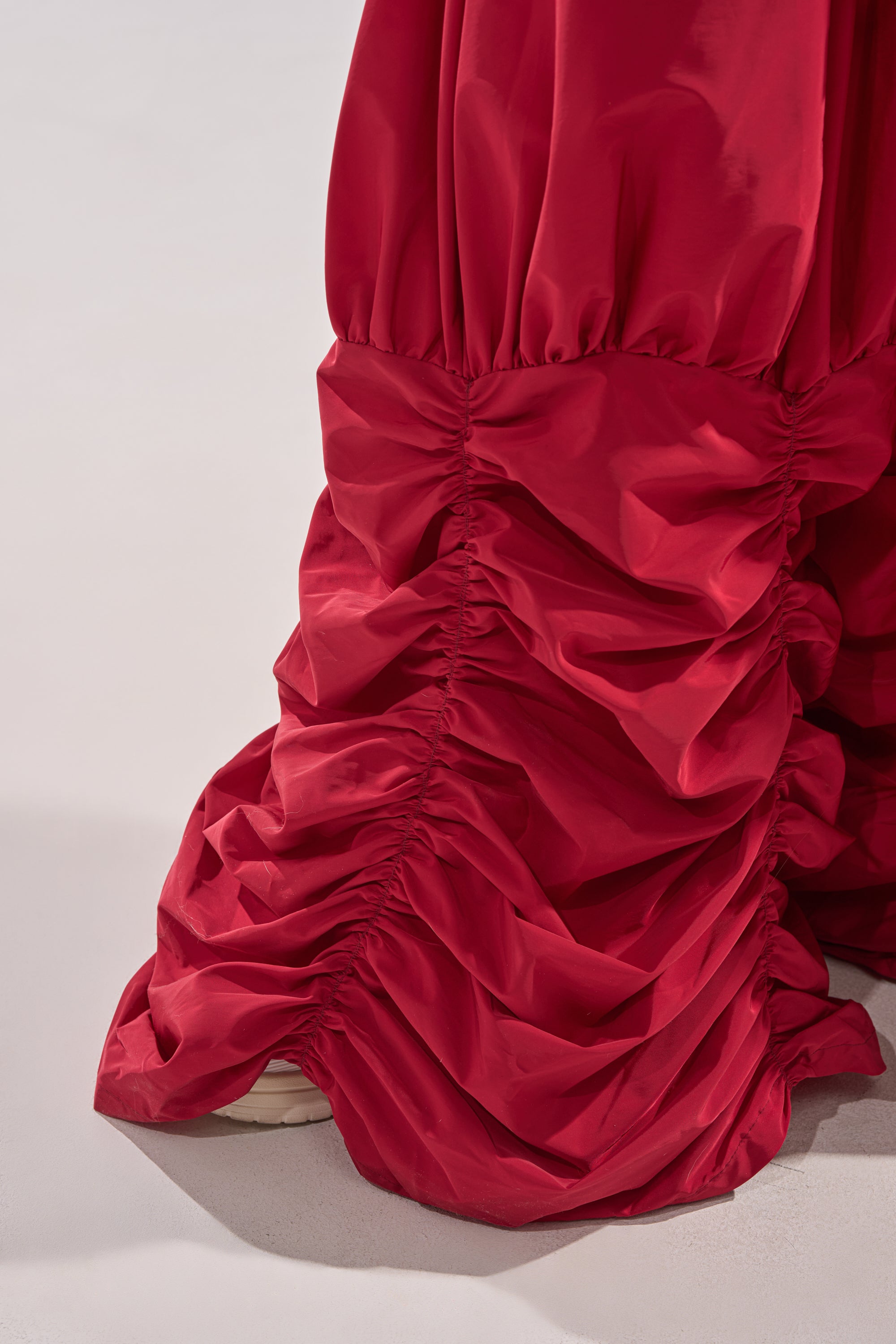 A close-up of the DON'T MIND ME STACKED JOGGER in red, featuring gathered, ruched fabric and voluminous folds reminiscent of parachute pants, partially covering a white shoe on a light surface. 8