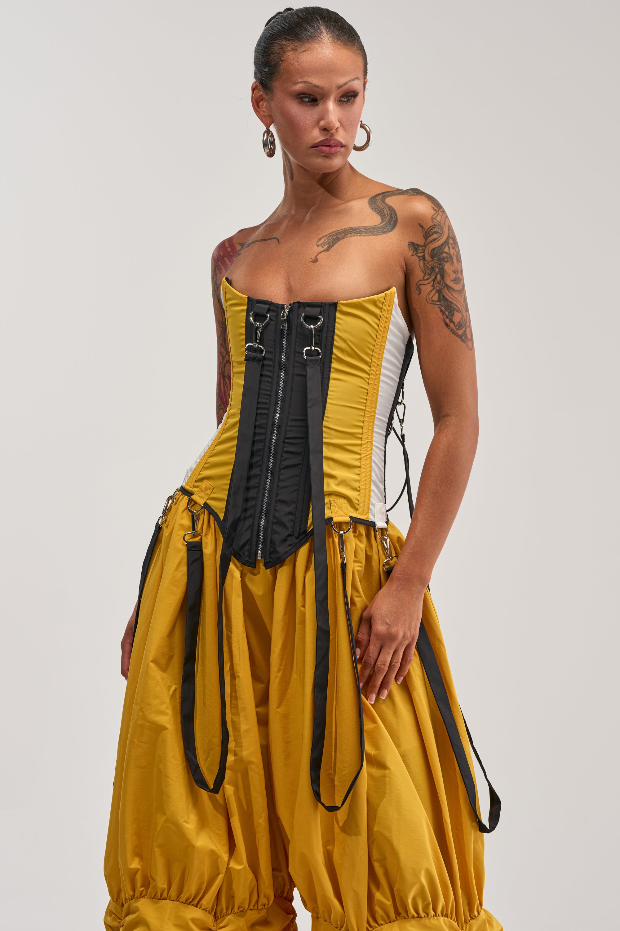 A tattooed woman with hoop earrings models the DON'T MIND ME CORSET IN YELLOW—a strapless, colorblocked piece with bold black front zippers—paired with a yellow balloon-style skirt featuring black straps, against a plain white background for an eye-catching streetwear look.