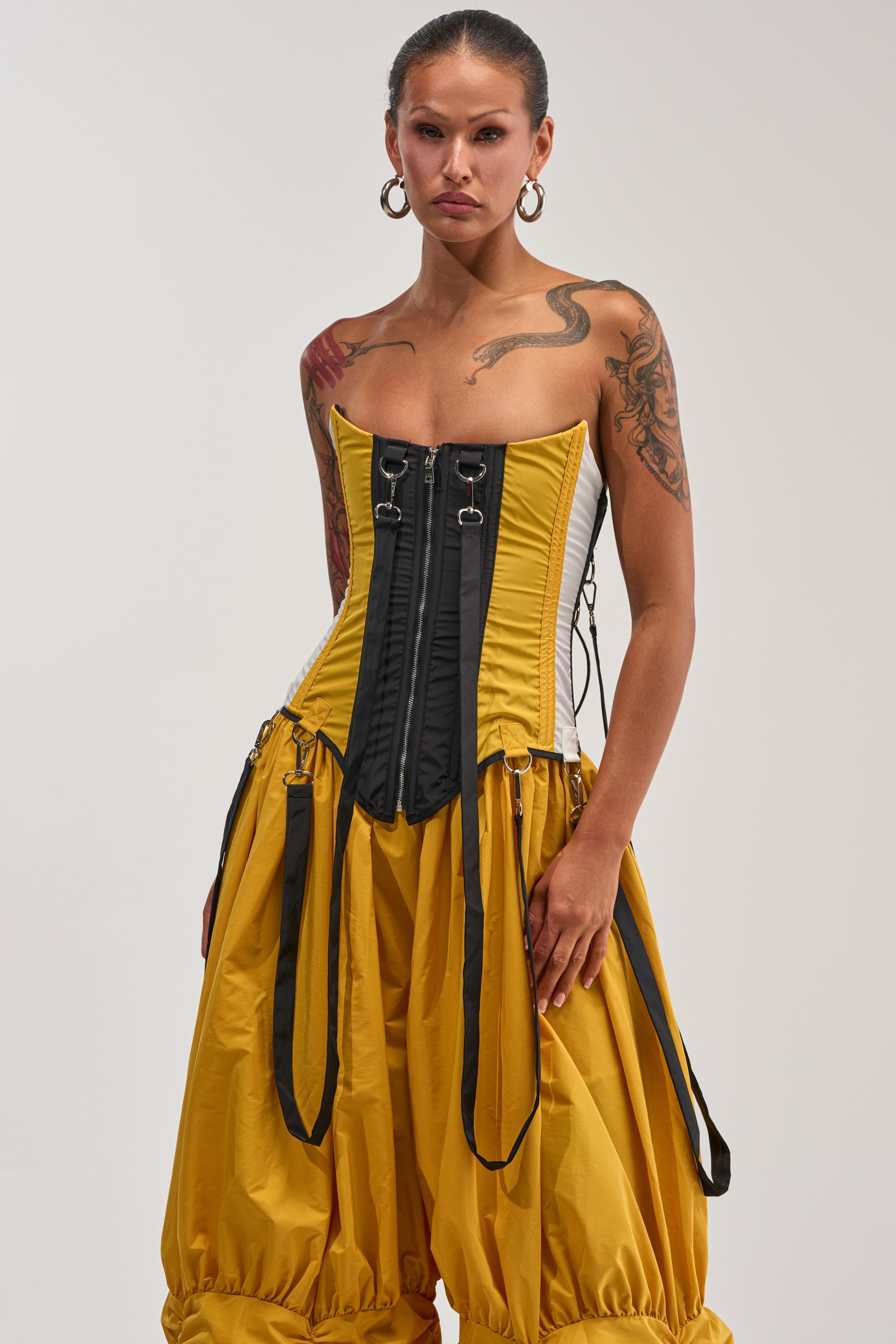Wearing the DON'T MIND ME CORSET IN YELLOW, a woman pairs the colorblocked yellow and black corset-style top with ballooned yellow pants, hoop earrings, and showcases prominent tattoos as she stands against a plain white background—embodying bold statement streetwear.