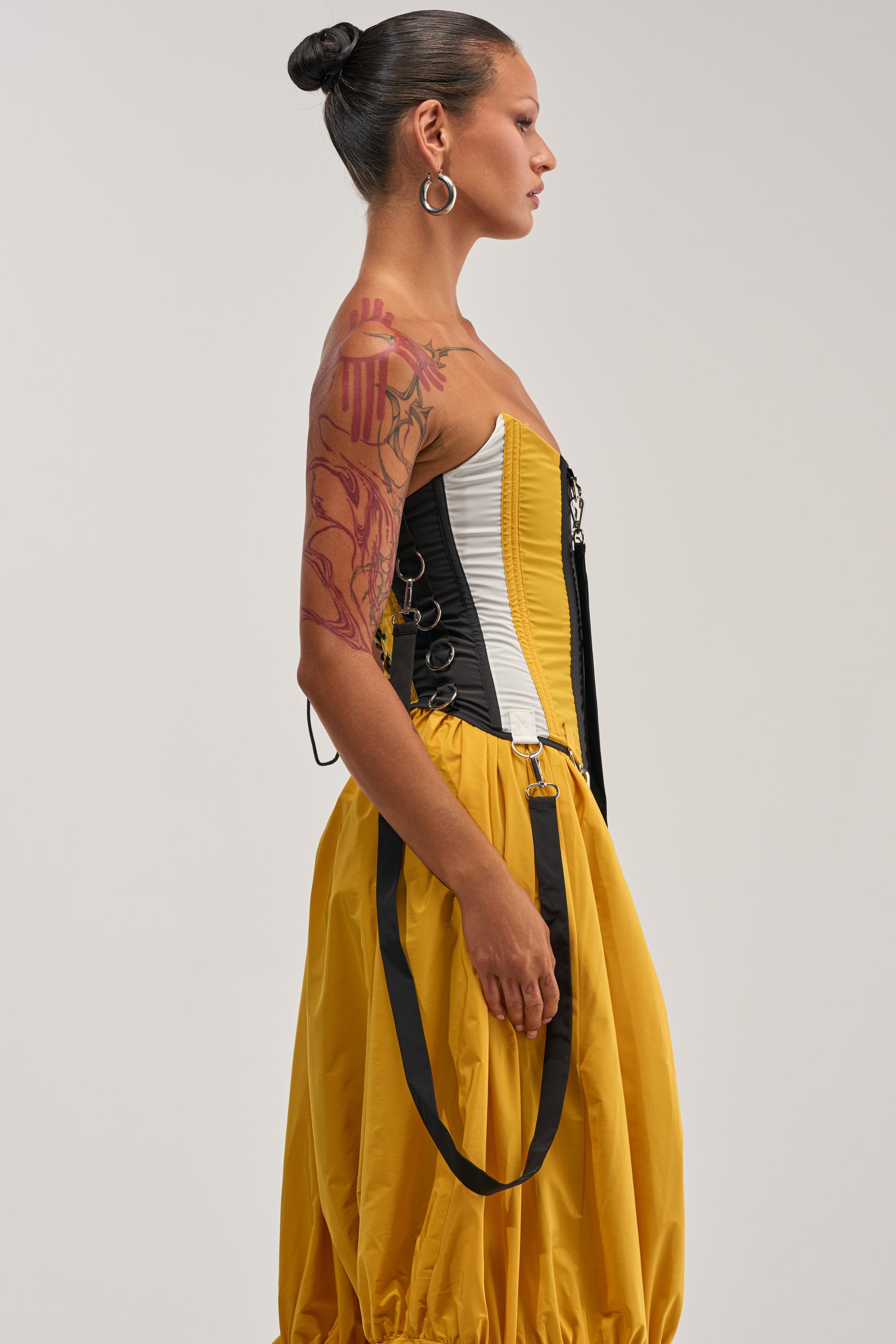 A person with a tattooed arm and hoop earrings stands in profile against a plain background, wearing the DON'T MIND ME CORSET IN YELLOW paired with a colorblocked skirt and black-and-white windbreaker details.