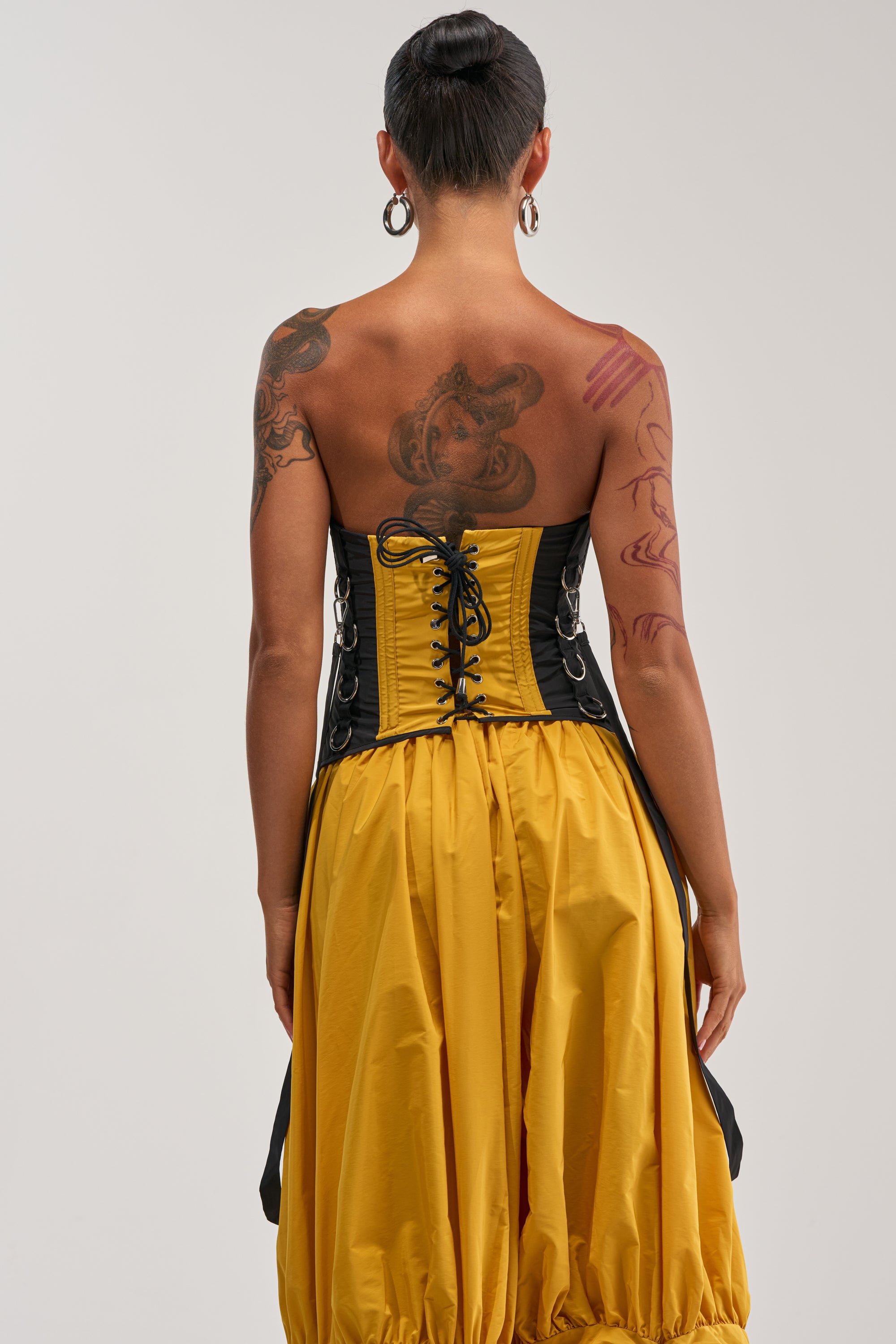 A person with tattoos, wearing the DON'T MIND ME CORSET IN YELLOW styled as a statement streetwear piece and a yellow skirt, stands facing away from the camera against a plain background.