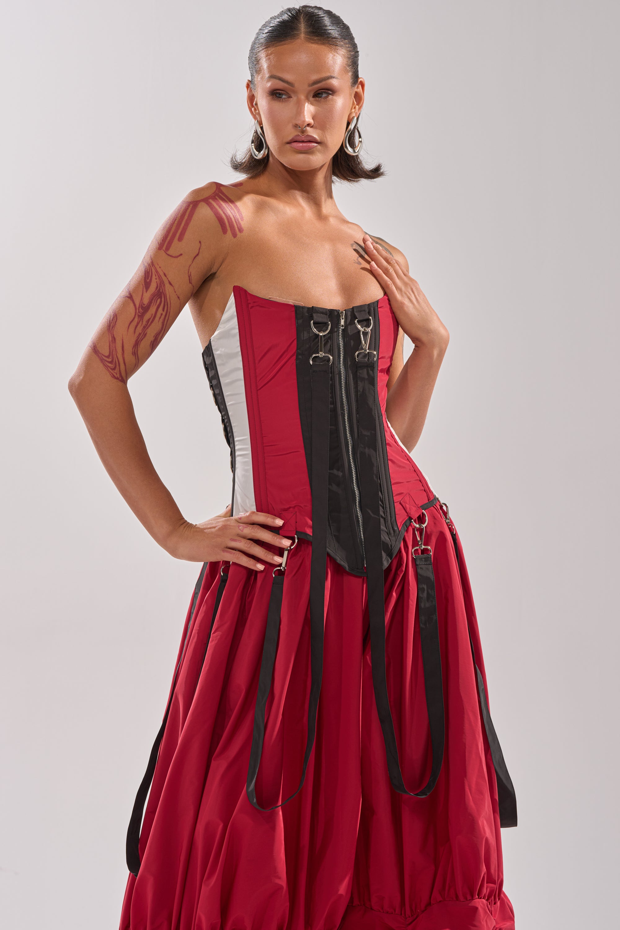A woman with hoop earrings and bold makeup poses confidently to the side, showcasing the DON'T MIND ME CORSET—a color-blocked dress featuring black straps, a voluminous red skirt, and visible tattoos on her arm. 0