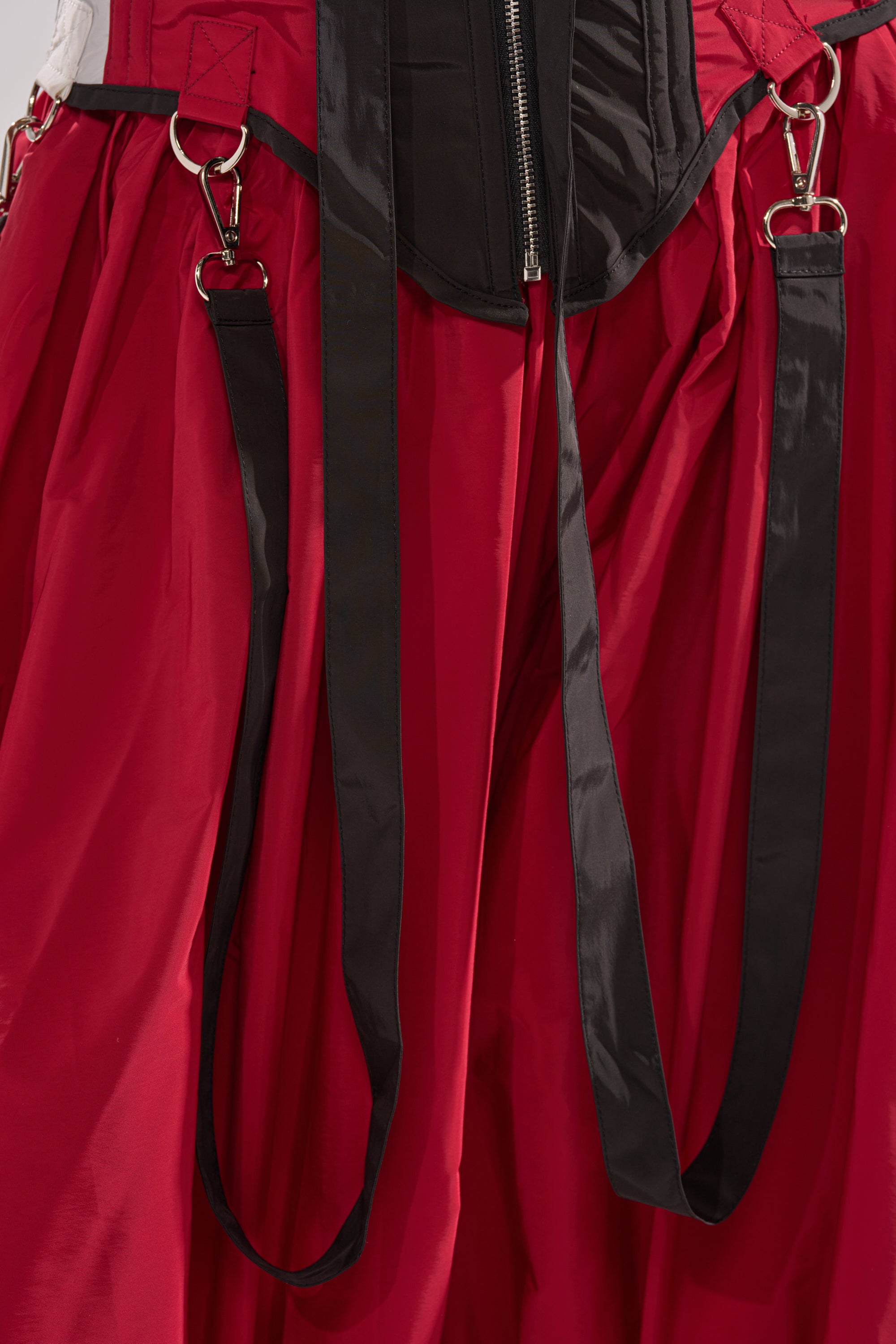Close-up of the DON'T MIND ME CORSET—red and black with black straps, silver metal clips, and a central black zipper. The smooth, slightly glossy material highlights the structured statement look. 13