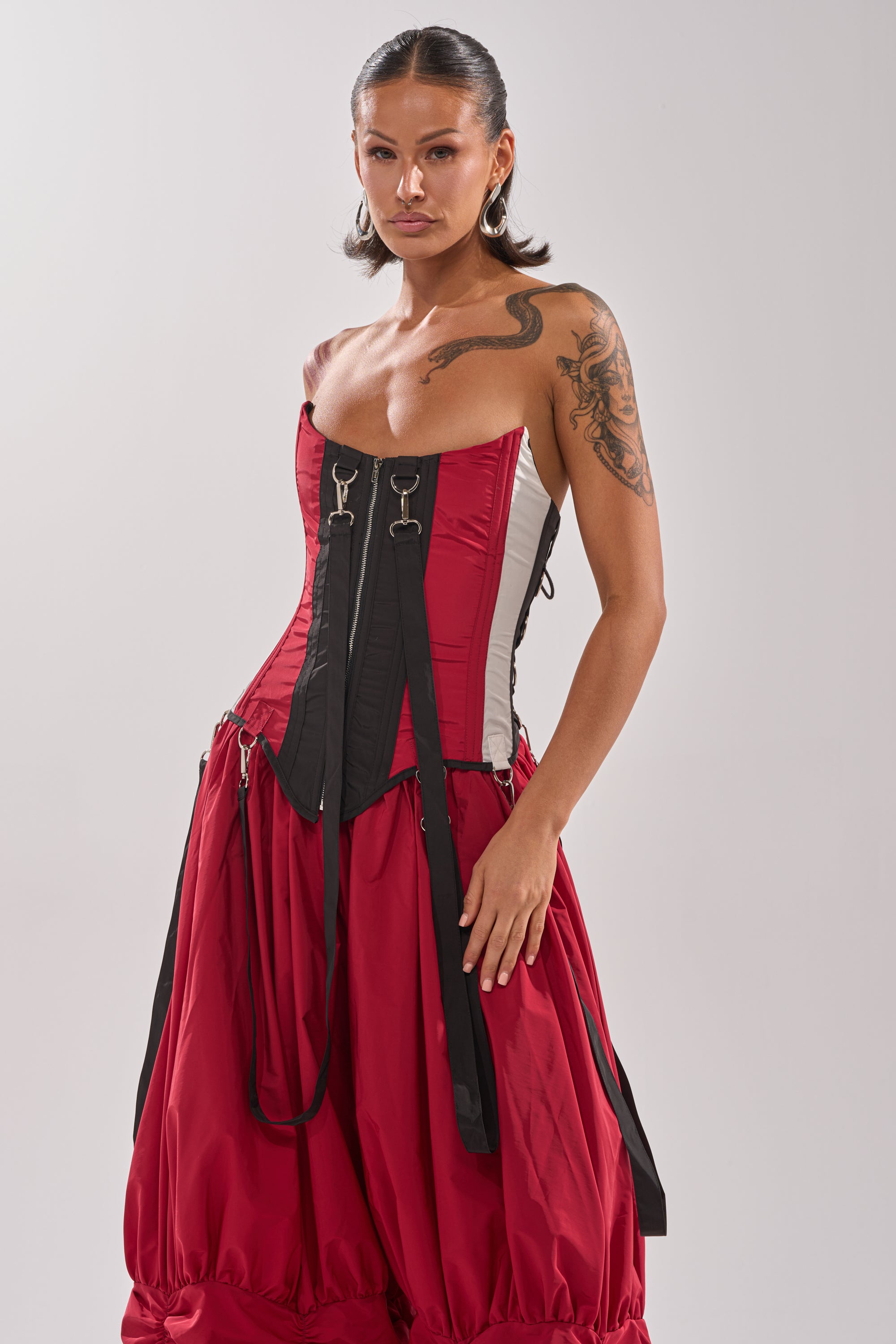 A woman with dark, slicked-back hair and a tattoo wears the DON'T MIND ME CORSET paired with a voluminous red-and-black color block skirt, standing confidently against a plain light background. 2