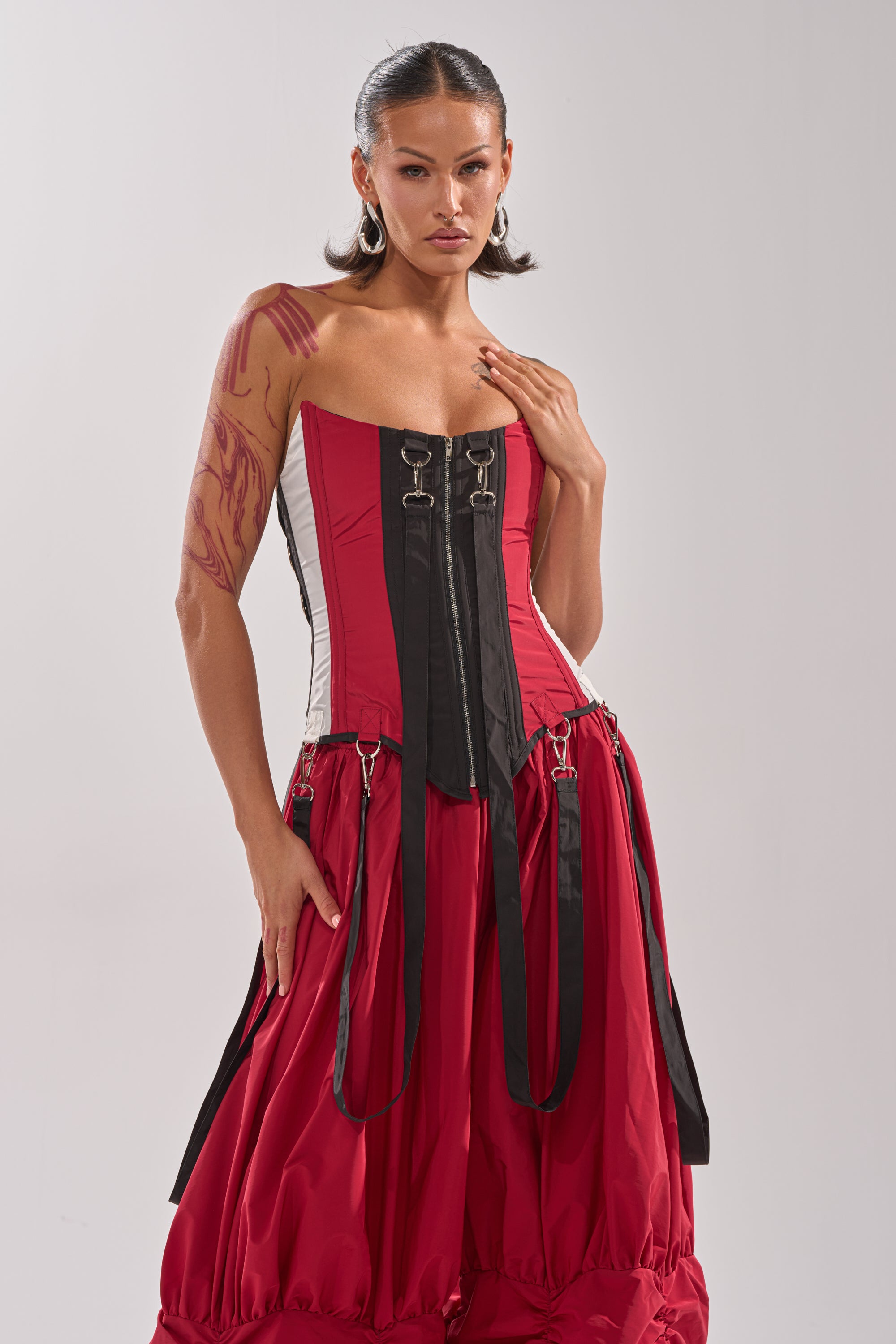 A woman wears the DON'T MIND ME CORSET—a bold red and black structured top with buckles and straps. With her hair slicked back, a large flame tattoo is visible on her right arm as she poses confidently against a plain background. 3