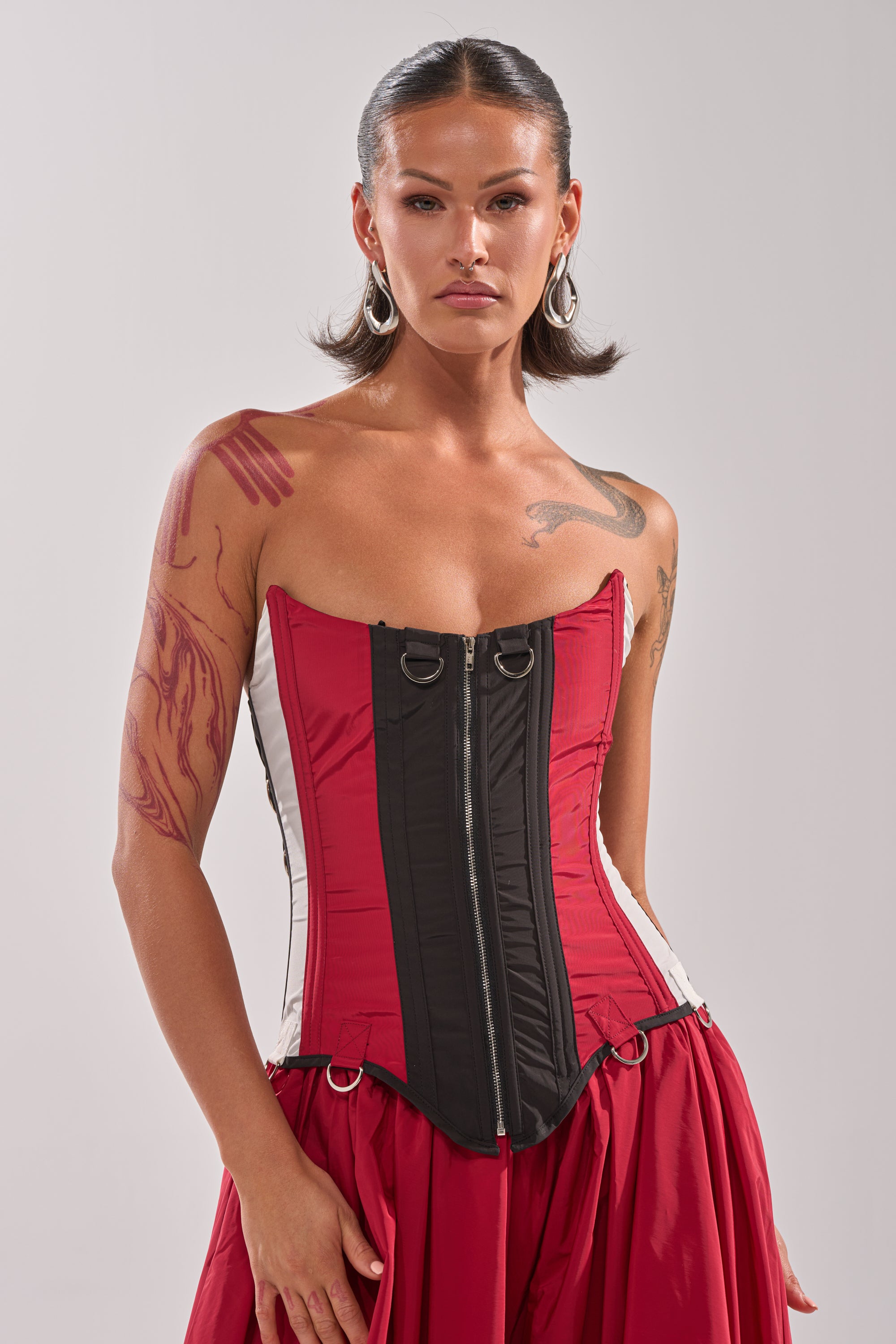 A person with slicked-back hair, hoop earrings, and visible tattoos models the DON'T MIND ME CORSET—a strapless red and black top—while standing against a light gray background. 4
