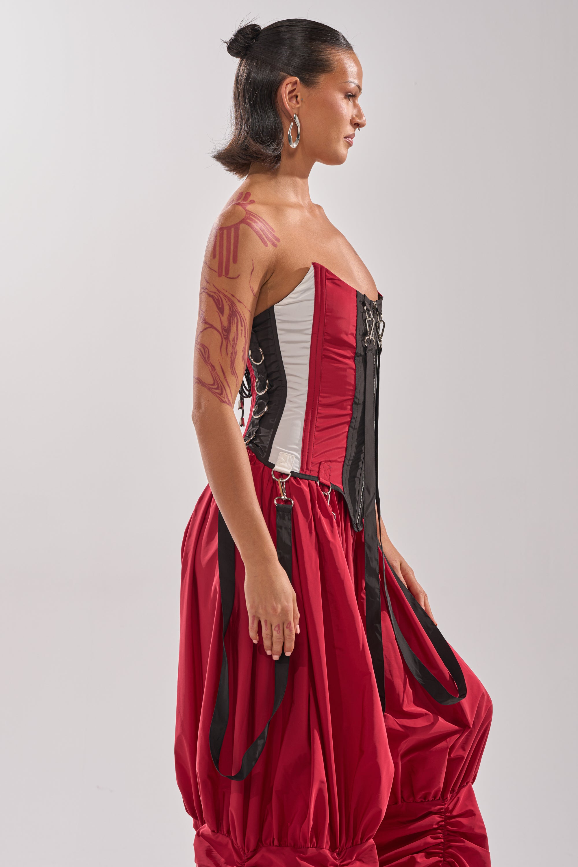 A woman in profile wears the DON'T MIND ME CORSET, a red-and-black color block top, paired with wide red pants. She has slicked-back short hair, large hoop earrings, and visible arm tattoos against a light background. 5
