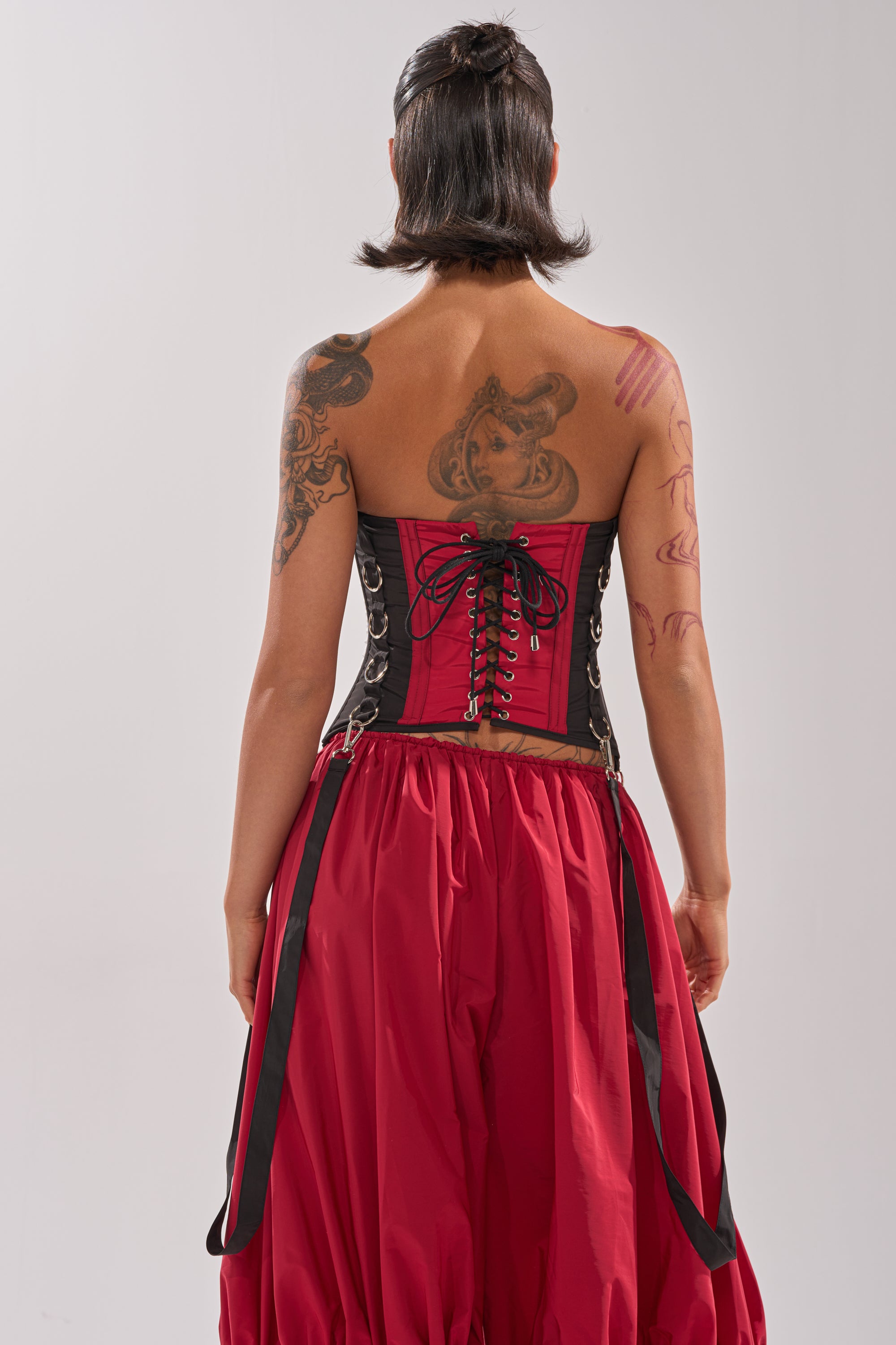 A person with short dark hair and visible tattoos models the DON'T MIND ME CORSET and a voluminous red skirt, standing with their back to the camera against a plain light background. 6