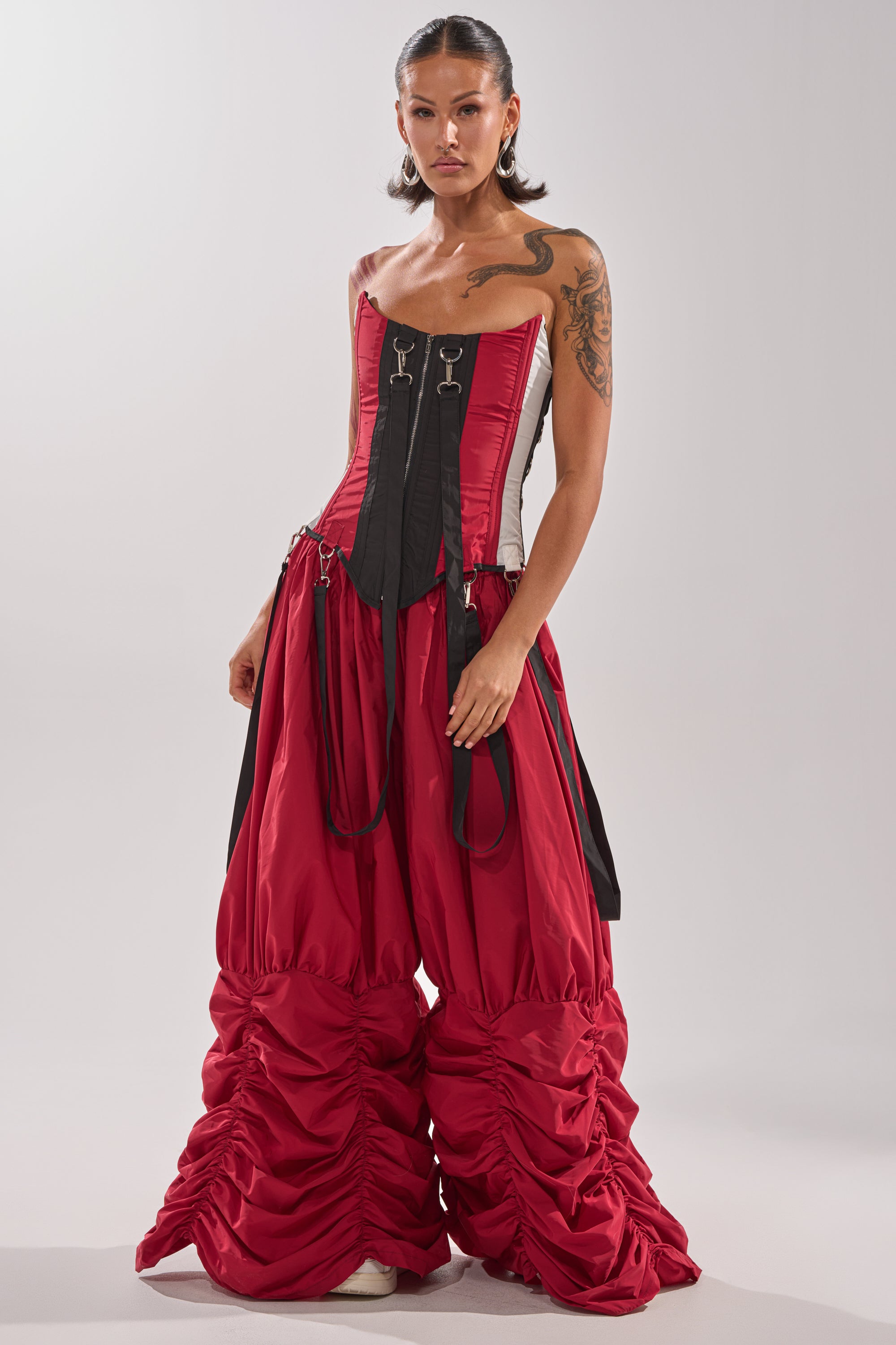 A woman confidently models the DON'T MIND ME CORSET, a structured statement top with decorative buckles, paired with voluminous ruched red pants against a light gray background. 8