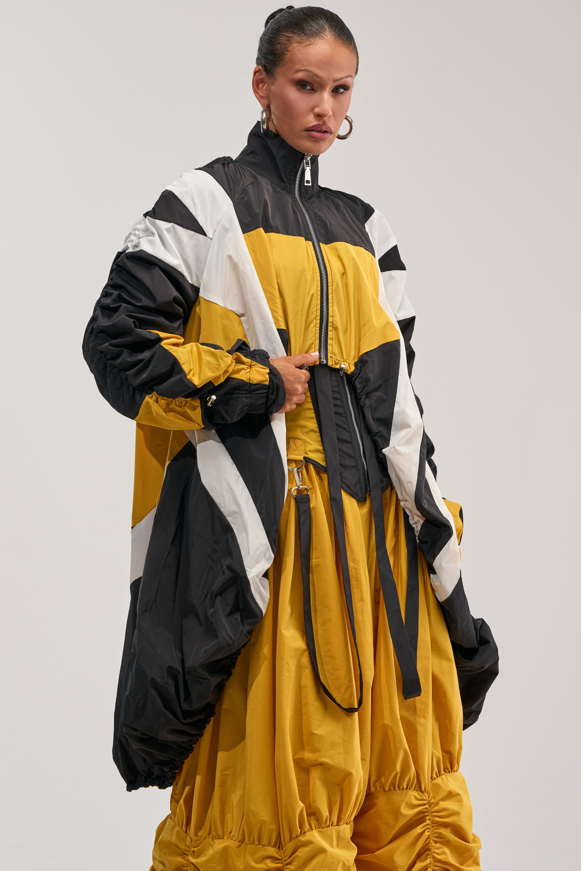 A woman wearing the DON'T MIND ME HIGH LOW WINDBREAKER IN YELLOW with matching yellow parachute-style pants, standing against a plain light background.