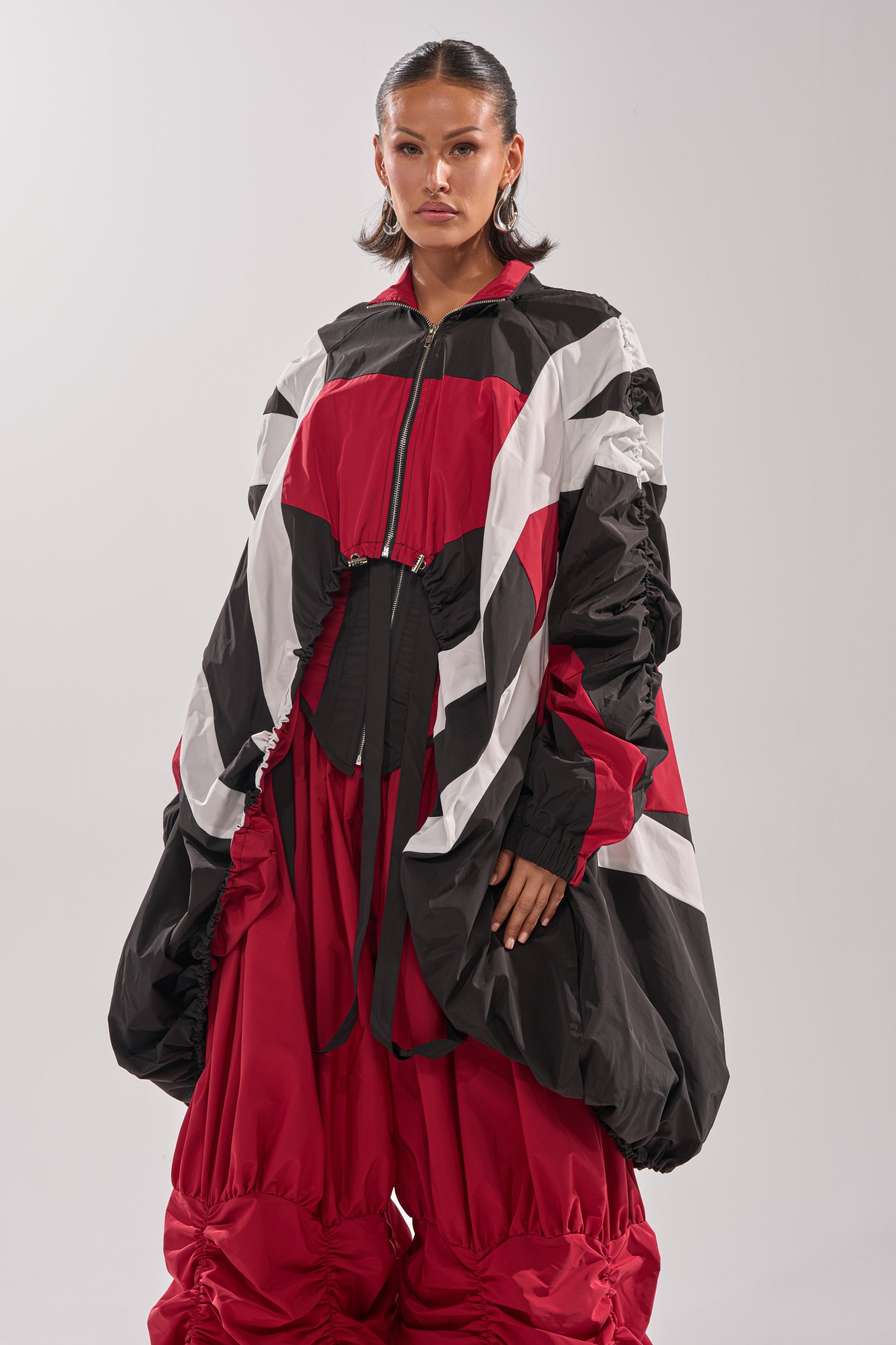 A woman wears the DON'T MIND ME HIGH LOW WINDBREAKER, an oversized, puffy jacket with bold black, white, and red geometric patterns, paired with matching wide ruched pants against a plain light background. 2