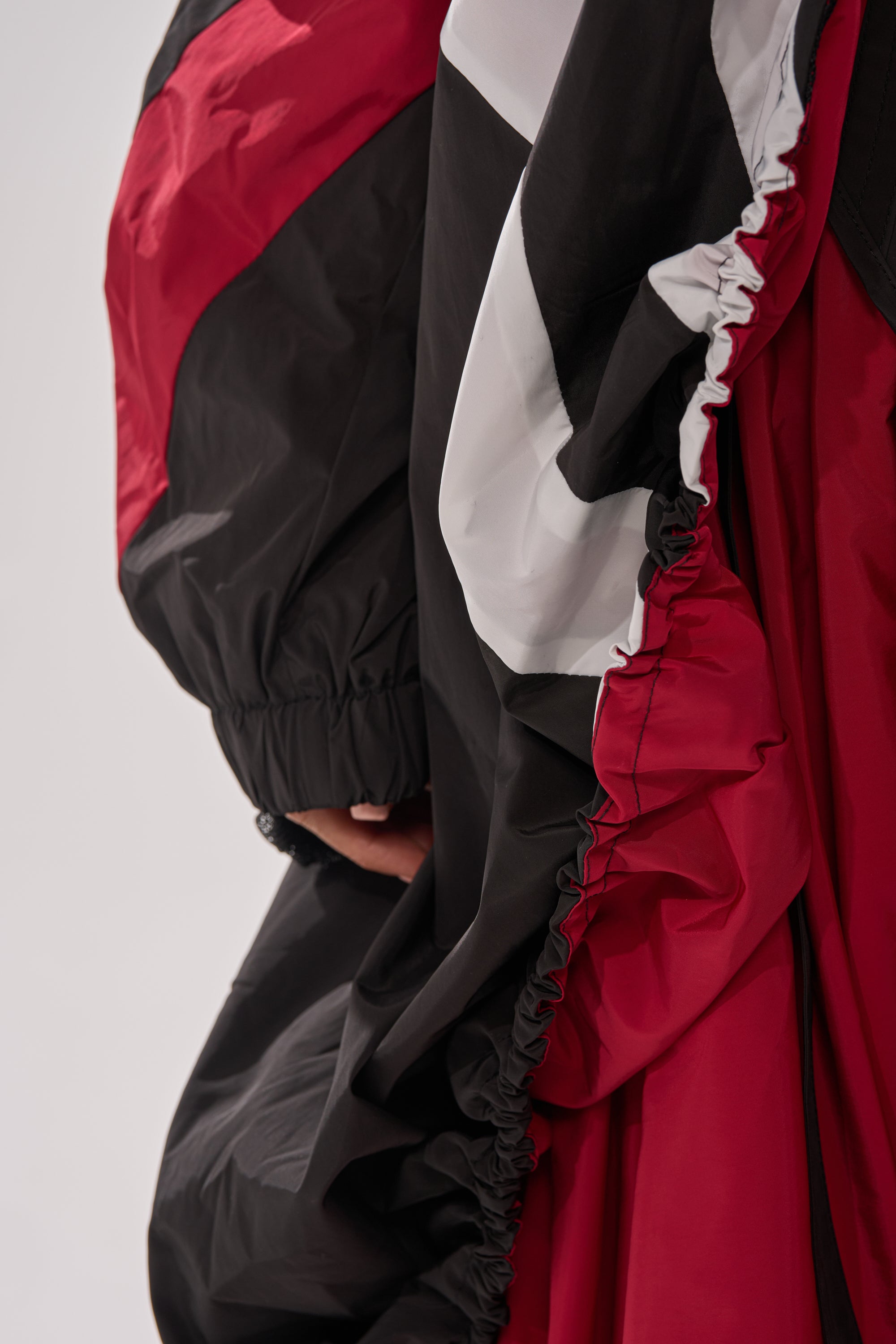 Close-up of a hand and part of an arm wearing the DON'T MIND ME HIGH LOW WINDBREAKER, a loose, color-blocked jacket in red, black, and white with ruched detailing. Background is plain and light-colored. 9