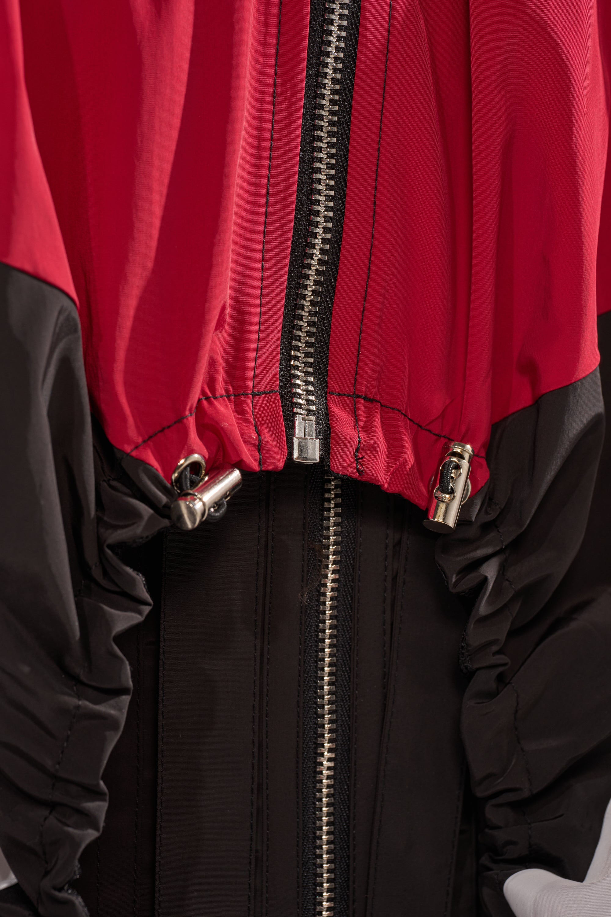 Close-up of the DON'T MIND ME HIGH LOW WINDBREAKER in red and black color-block, featuring a silver center zipper and adjustable drawstring toggles at the hem on each side. 10