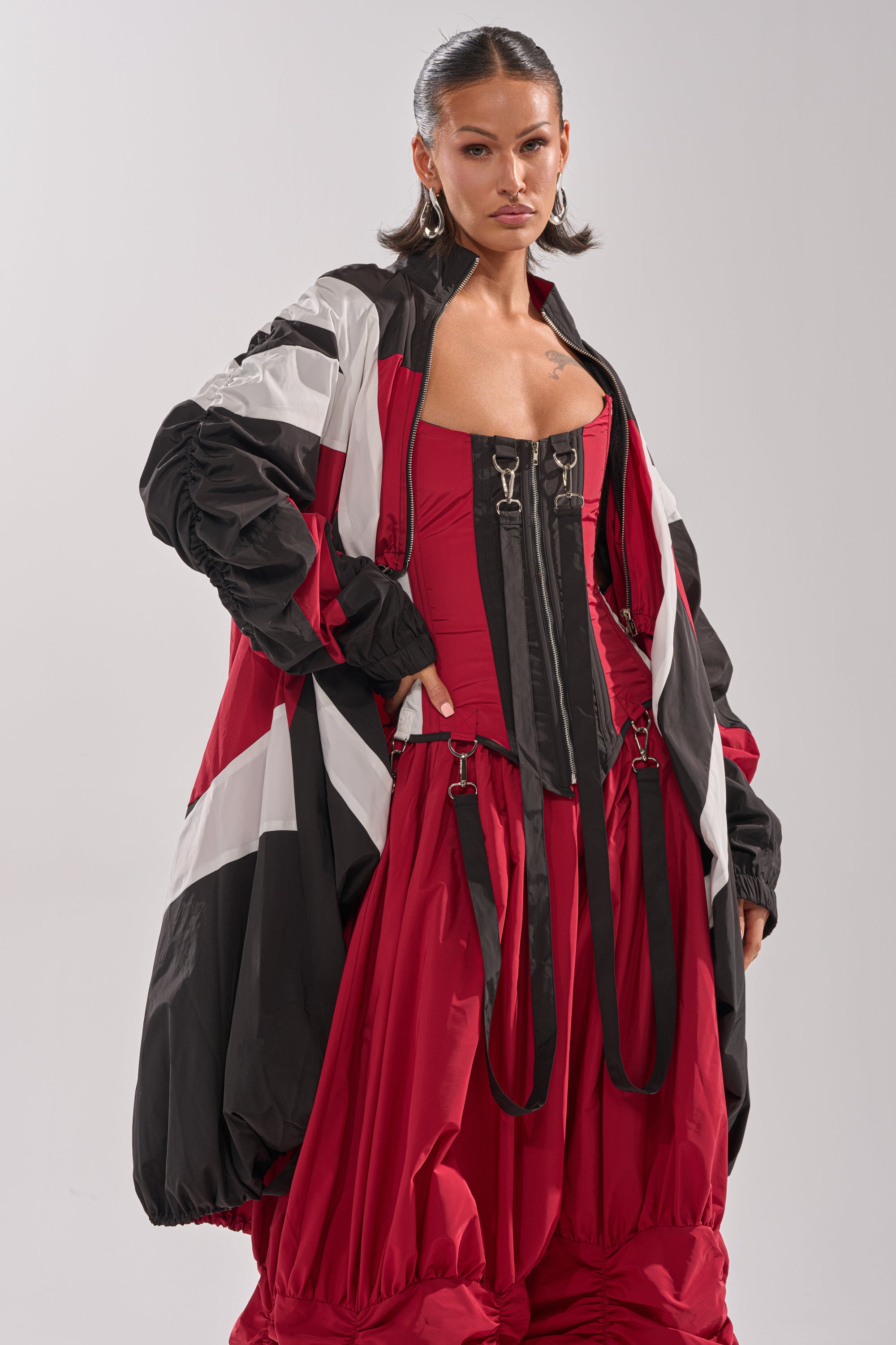 A woman exudes confidence in the DON'T MIND ME HIGH LOW WINDBREAKER, styled over a red corset top with buckles and paired with striking red pants featuring black strap details. 3
