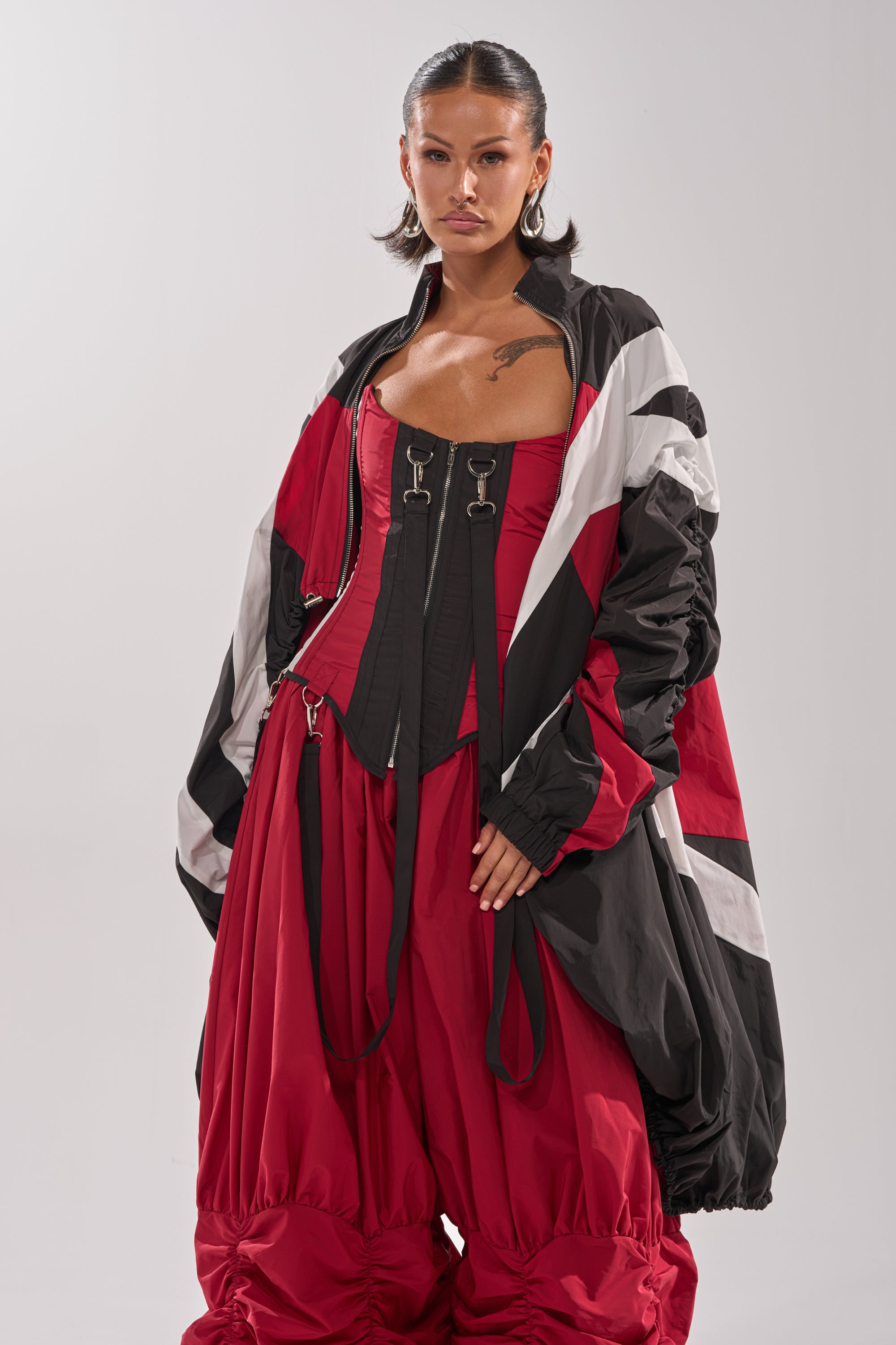 A woman with slicked-back hair and hoop earrings poses against a light background in the DON'T MIND ME HIGH LOW WINDBREAKER with white stripes, a black buckle corset, and bold red pants. 4
