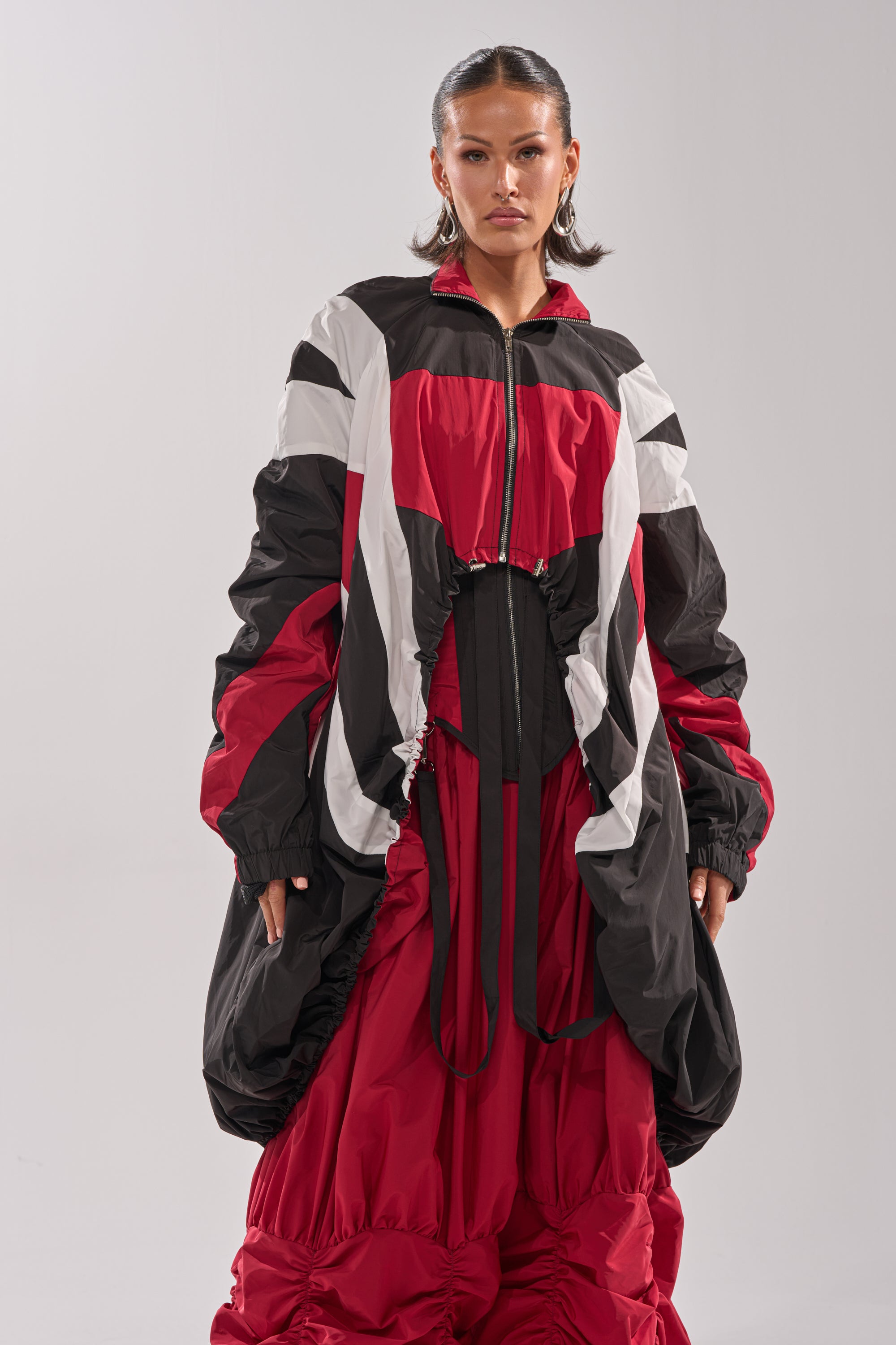 Wearing the DON'T MIND ME HIGH LOW WINDBREAKER, a woman pairs its bold, oversized color-block design with a voluminous red skirt. She stands before a plain light background and gazes confidently at the camera. 5
