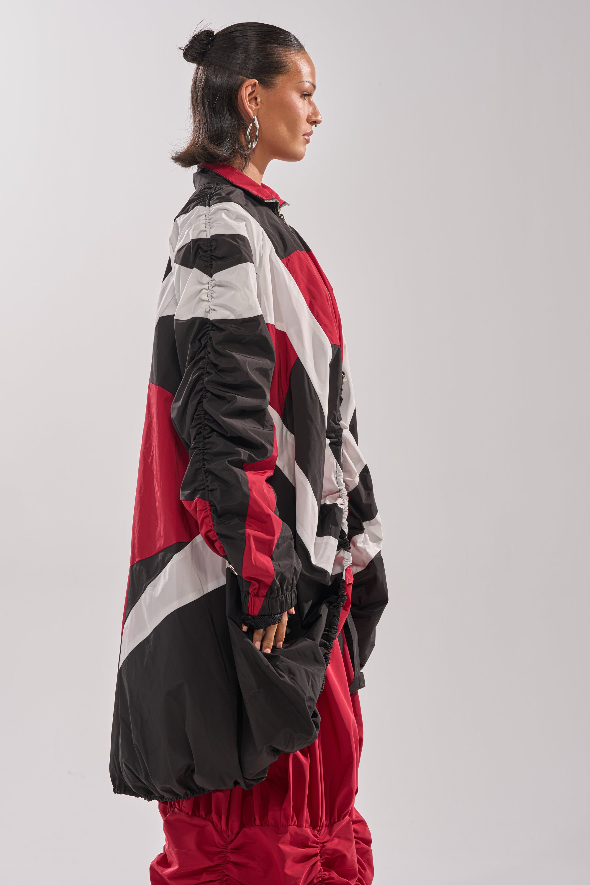 A person with slicked-back hair poses in profile against a plain light background, wearing the DON'T MIND ME HIGH LOW WINDBREAKER—an oversized color-block jacket and pants with bold red, black, and white geometric patterns. 6