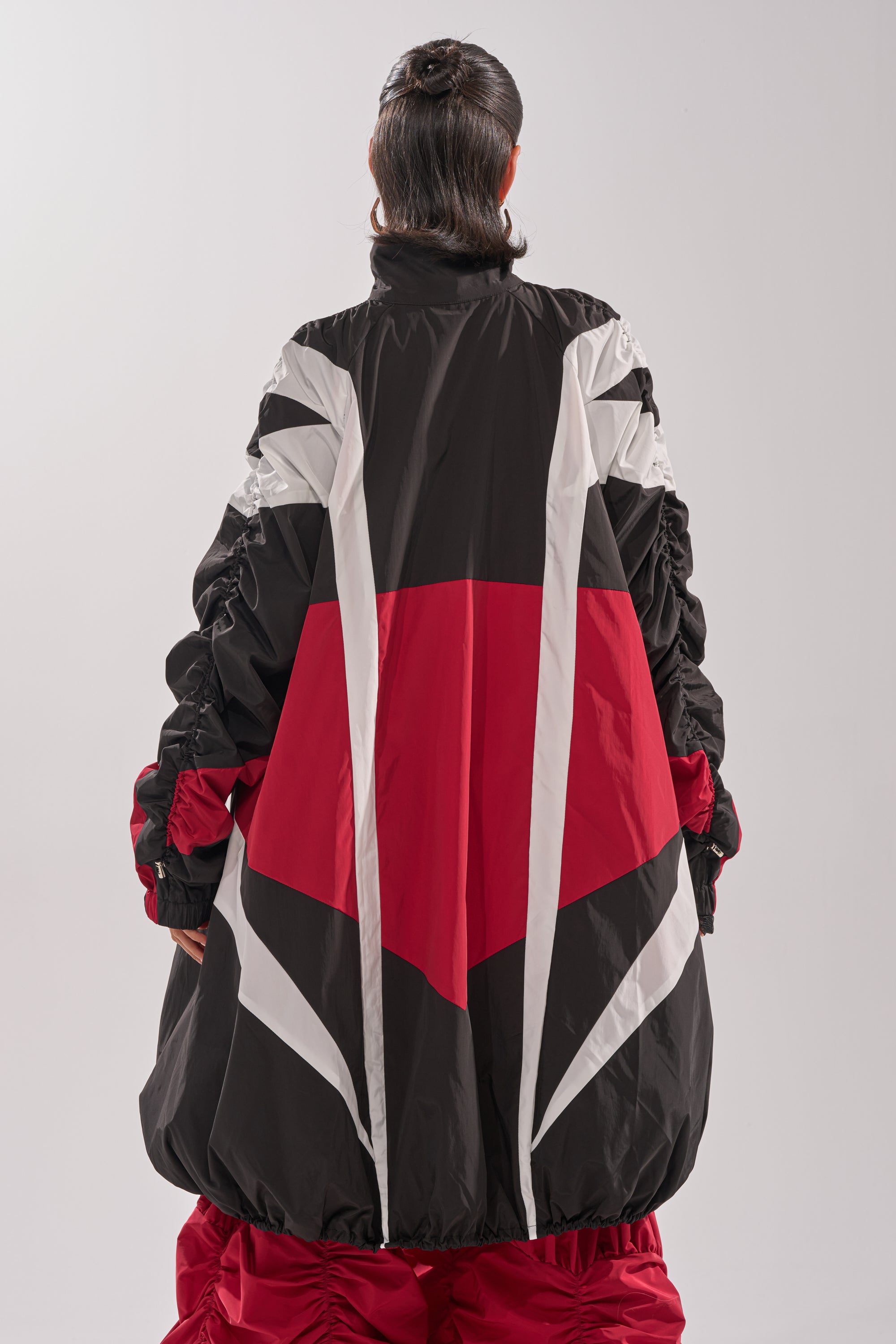 A person with long dark hair faces away, wearing the DON'T MIND ME HIGH LOW WINDBREAKER—an oversized jacket featuring bold red, black, and white geometric color-block patterns. The background is plain and light-colored. 7