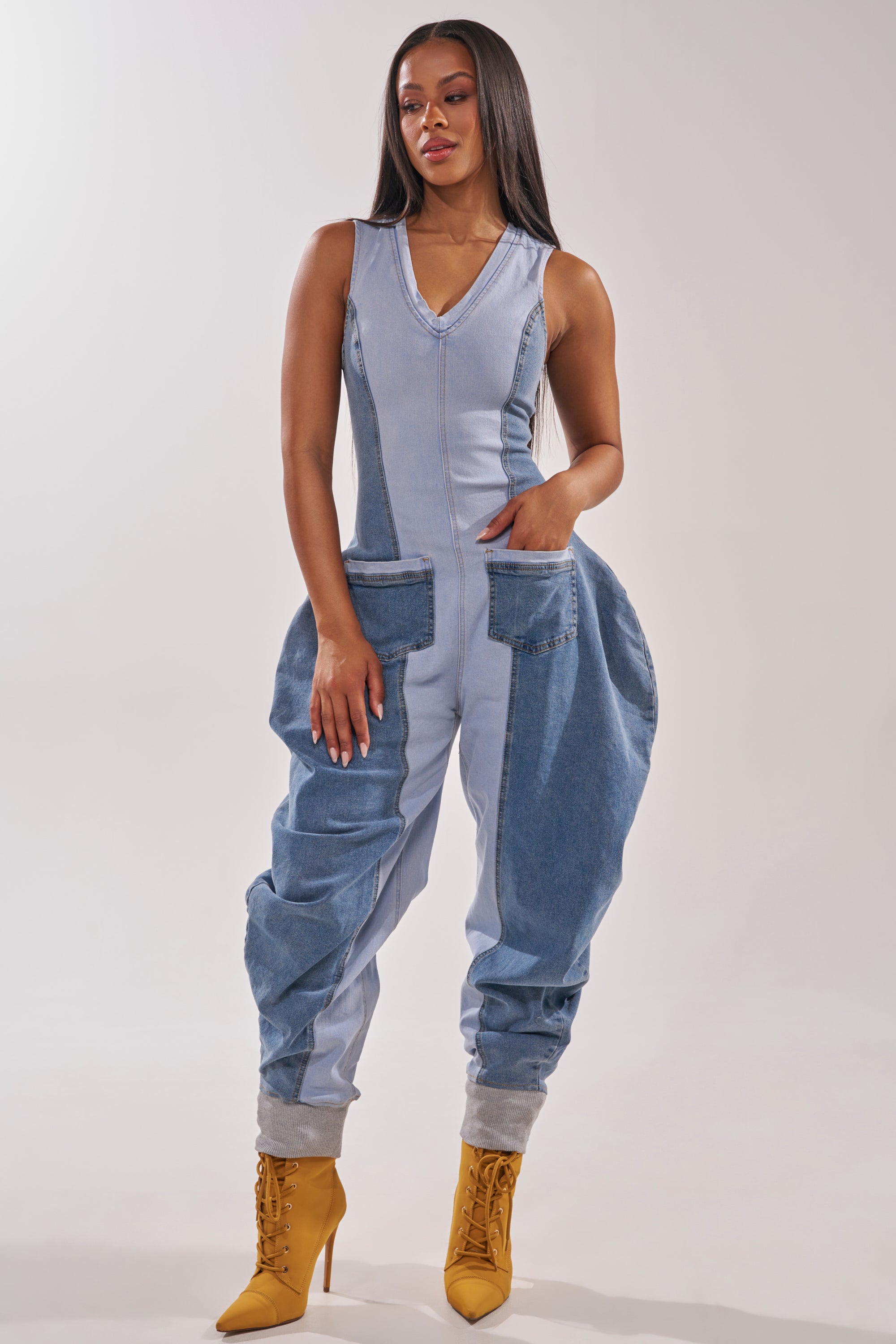 A woman faces forward in the TALK OF THE STREET DENIM JUMPSUIT, a sleeveless, two-tone denim piece with large rounded pockets and loose legs. She pairs it with mustard yellow ankle boots against a plain, light background. 0