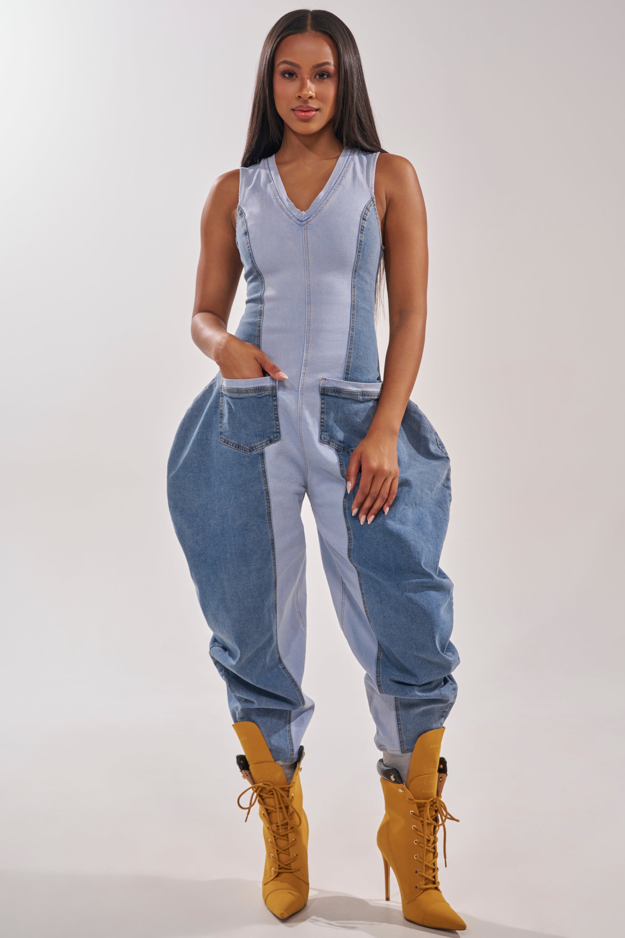 A woman faces forward in the TALK OF THE STREET DENIM JUMPSUIT, featuring large side pockets and wide legs, styled with mustard yellow lace-up heeled boots. The background is plain and light-colored. 1