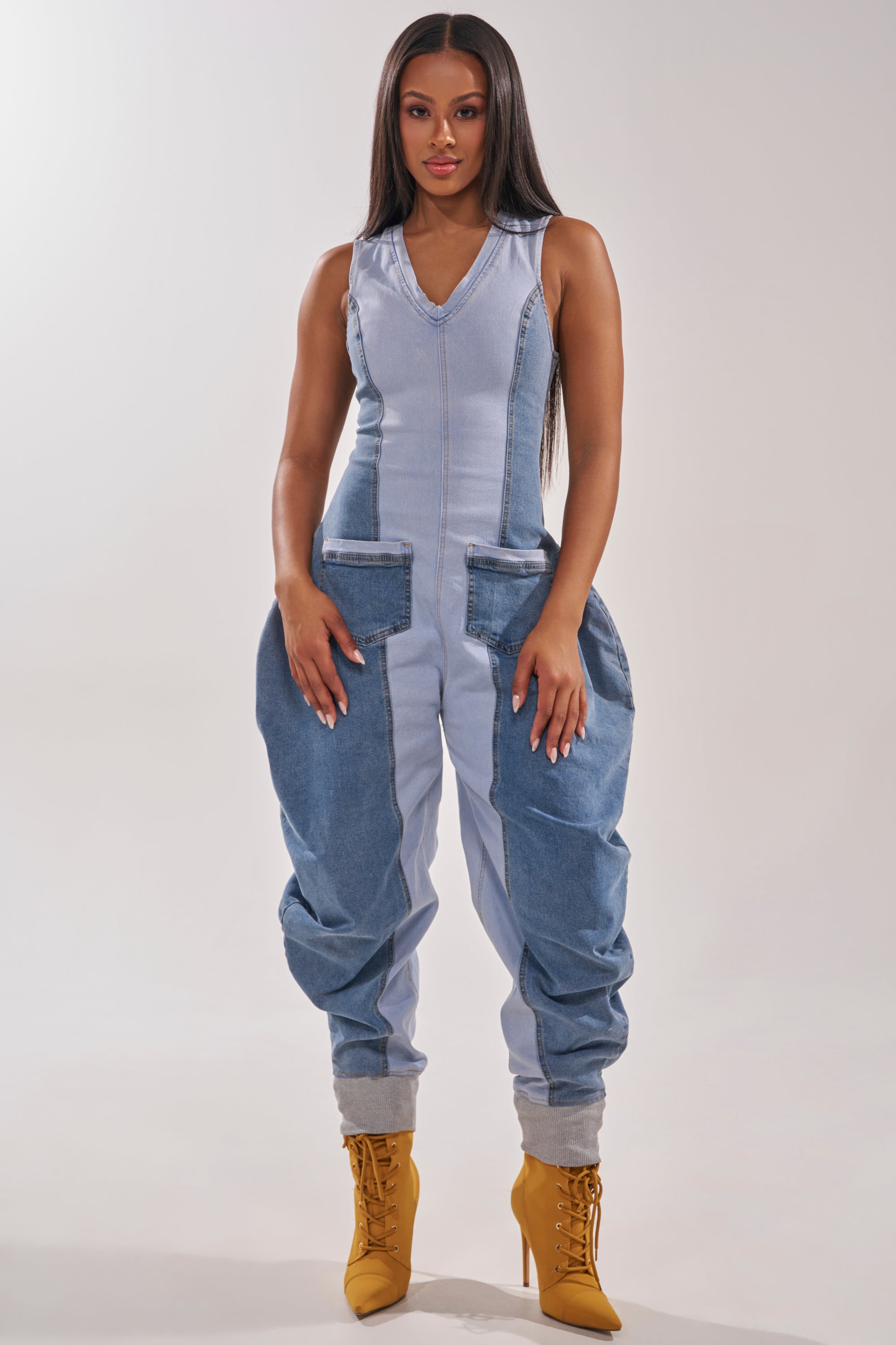 A woman faces forward in the TALK OF THE STREET DENIM JUMPSUIT, a utility-inspired, sleeveless piece with large side pockets and loose legs, styled with tan high-heeled boots against a plain, neutral background. 2