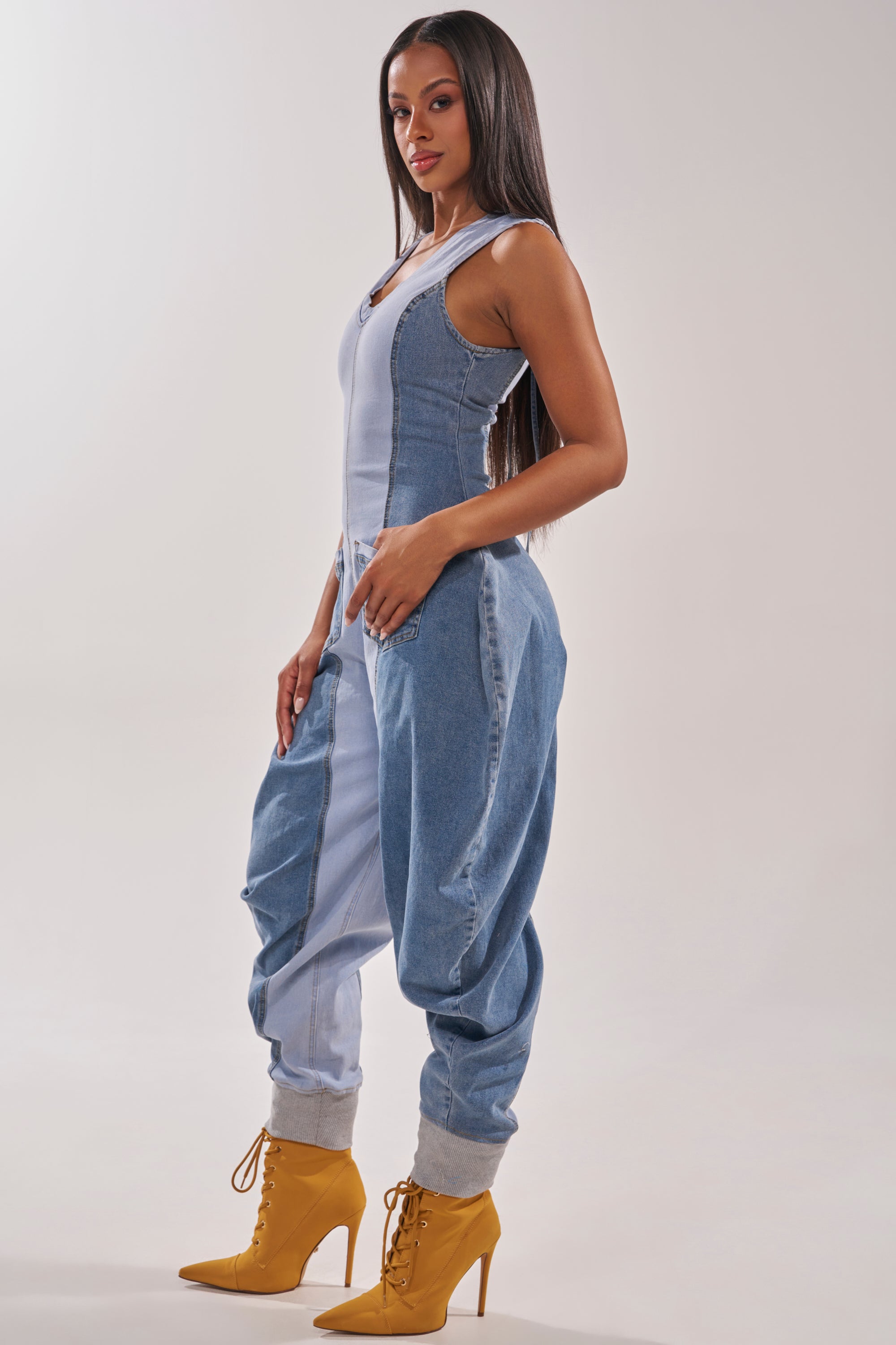 A woman with long straight hair poses sideways in the TALK OF THE STREET DENIM JUMPSUIT, a utility-inspired, sleeveless wide-leg style, paired with yellow lace-up high heel boots for a modern streetwear look against a light background. 3