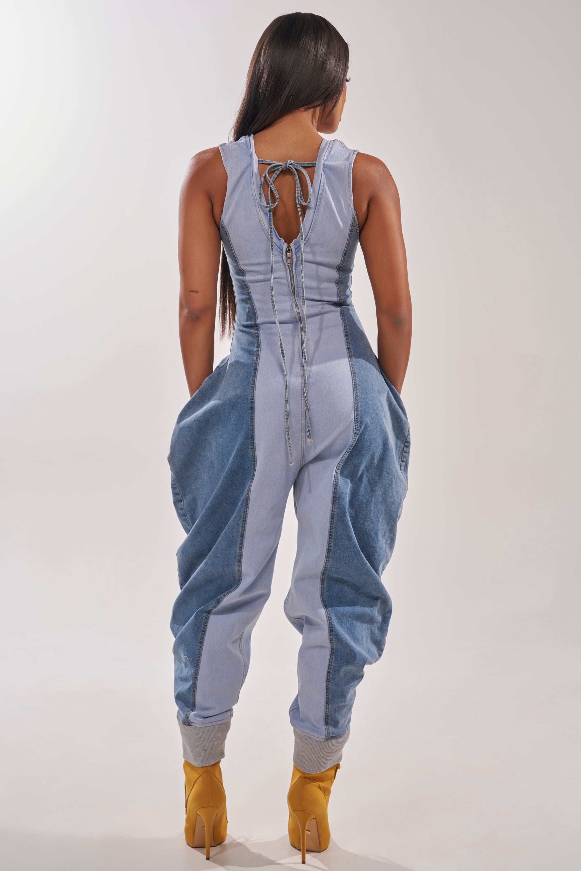 A woman with long dark hair faces away, wearing the TALK OF THE STREET DENIM JUMPSUIT featuring patchwork, a lace-up back, loose legs, and cuffed ankles. She stands against a neutral background with yellow heeled boots. 4