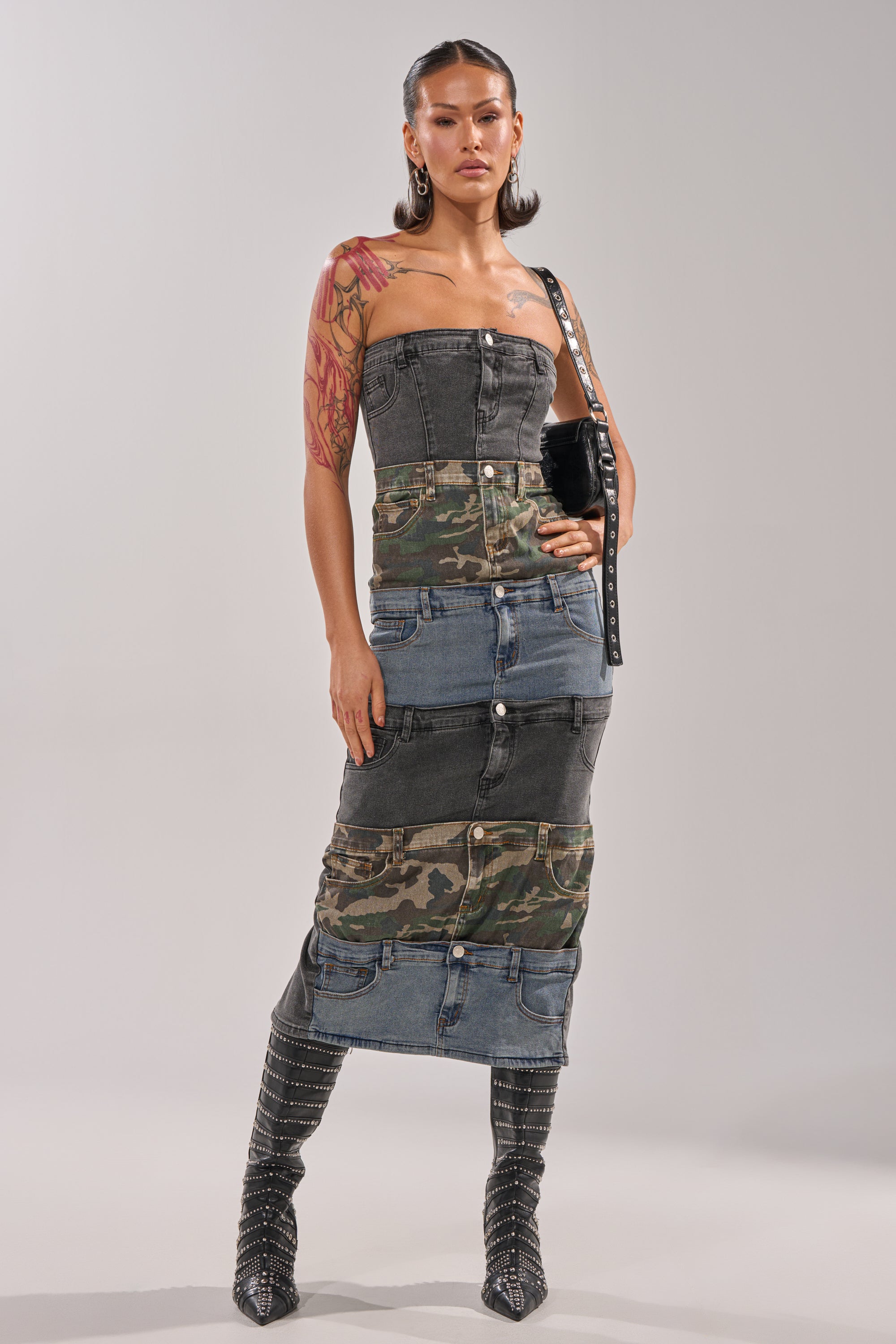A woman stands against a light background in the PATCHWORK PARADOX DENIM MAXI DRESS, featuring camouflage details. She pairs it with black studded boots, a black purse, large earrings, and displays a bold red tattoo on her right arm. 0