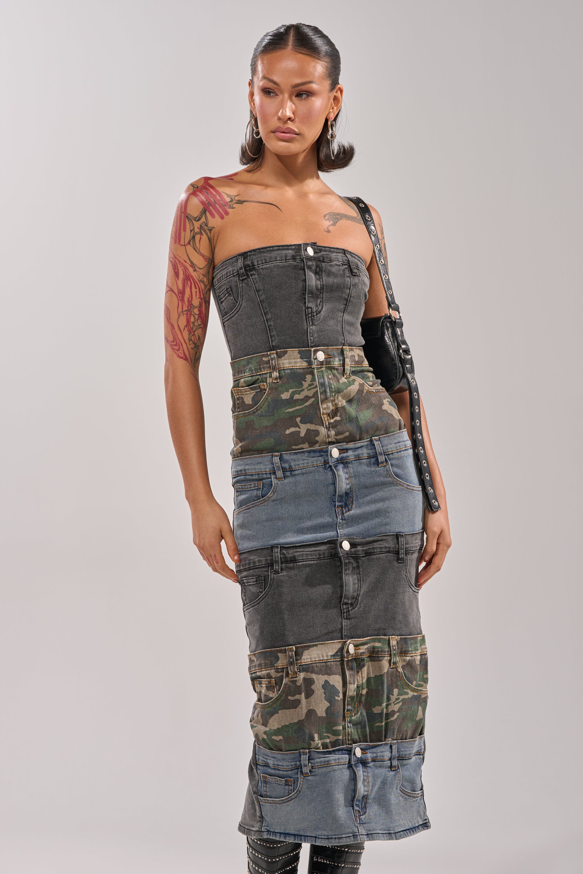 A woman showcases street style in the PATCHWORK PARADOX DENIM MAXI DRESS, a strapless patchwork denim piece with camo contrast, large buttons, and pocket details. She accessorizes with hoop earrings, an arm tattoo, and a black purse. 2