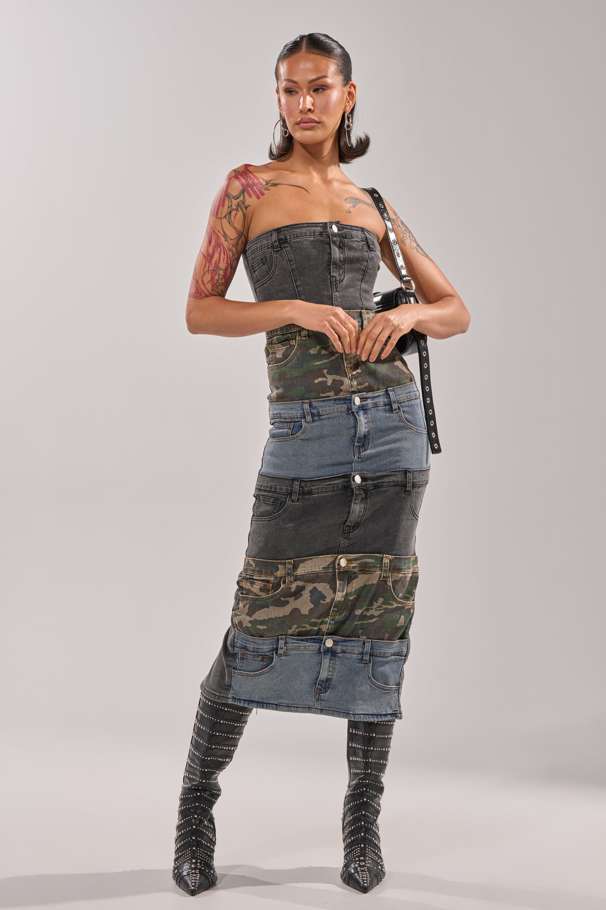A woman exudes bold street style in the PATCHWORK PARADOX DENIM MAXI DRESS with camo contrast and button details, styled with studded knee-high boots, a black belt, hoop earrings, and visible tattoos. 5