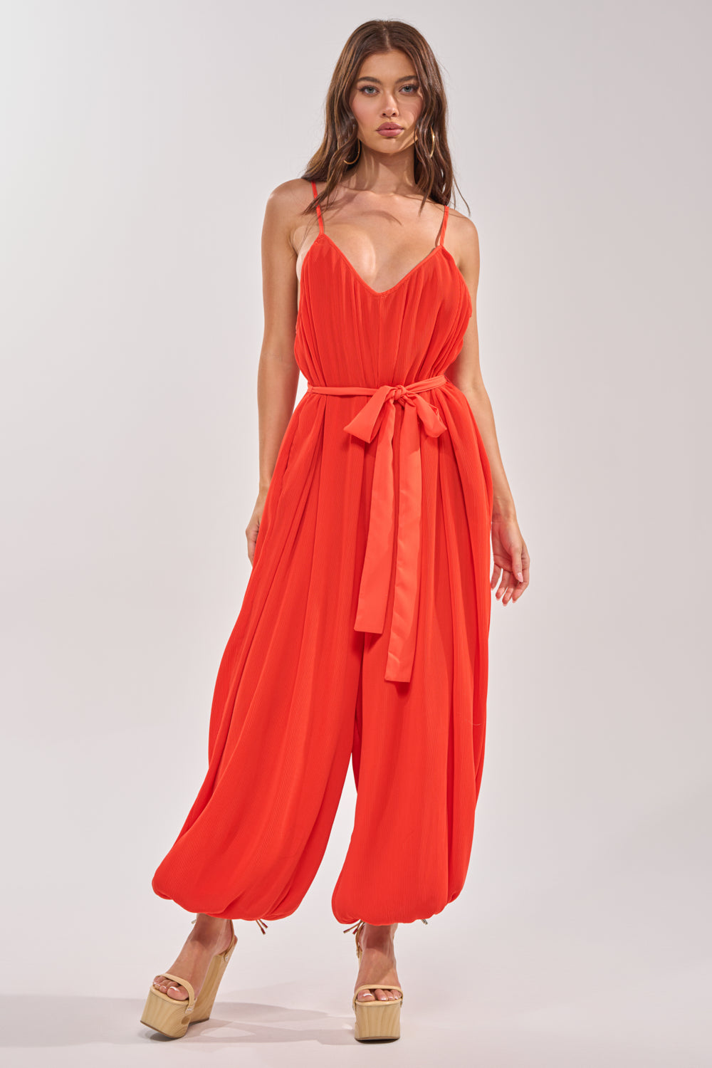 A woman wears the VACATIONING PLEATED JUMPSUIT IN ORANGE, featuring vibrant pleats, wide legs, a belt, and spaghetti straps. Paired with beige platform sandals, she stands against a plain light background for a perfect vacation look. 0