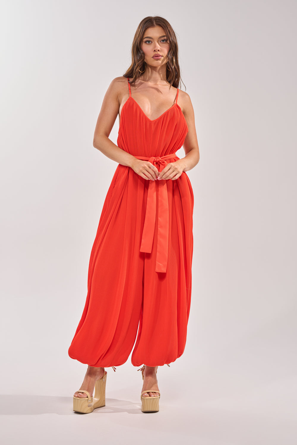 A woman wears the VACATIONING PLEATED JUMPSUIT IN ORANGE with a tied waist and ankle cuffs, paired with beige platform heels, standing against a plain light background—an ideal wide-leg jumpsuit for vacation style. 1